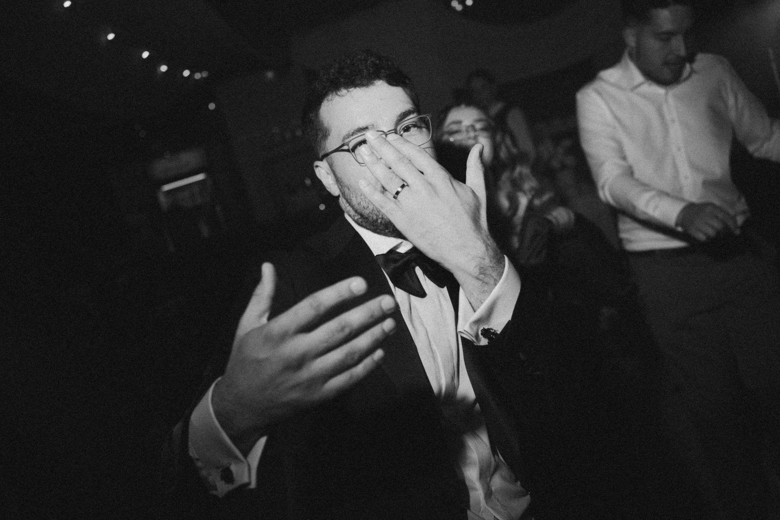 Black and white photo of a young man in a tuxedo and bowtie, wearing glasses, smiling and covering his mouth with his hand, at a social event with other people in the background.