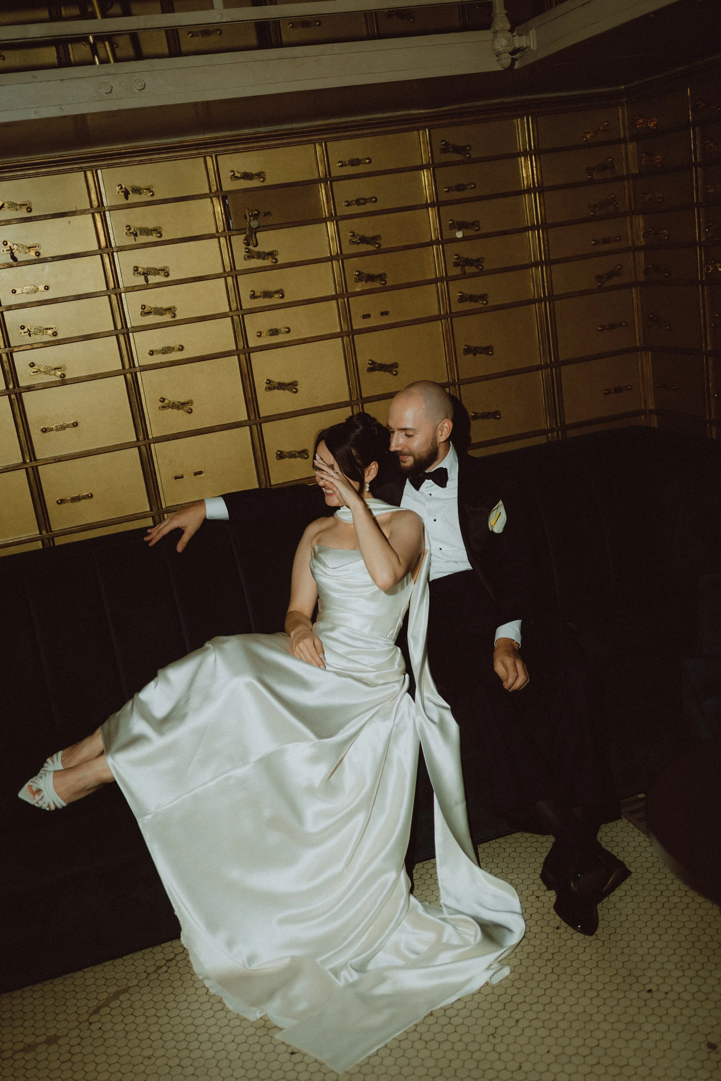 A man and woman dressed in formal wedding attire sitting on a black bench, with the woman covering her face and the man smiling, in front of a wall of mailboxes.