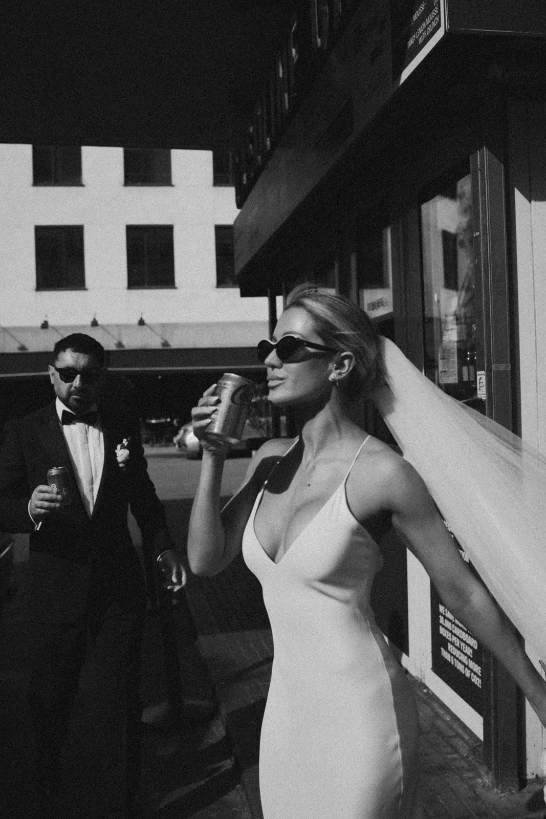 A woman in a white dress with sunglasses holding a can of beverage near a door, with a man in a tuxedo and dark sunglasses standing nearby, both in an urban setting with buildings in the background.