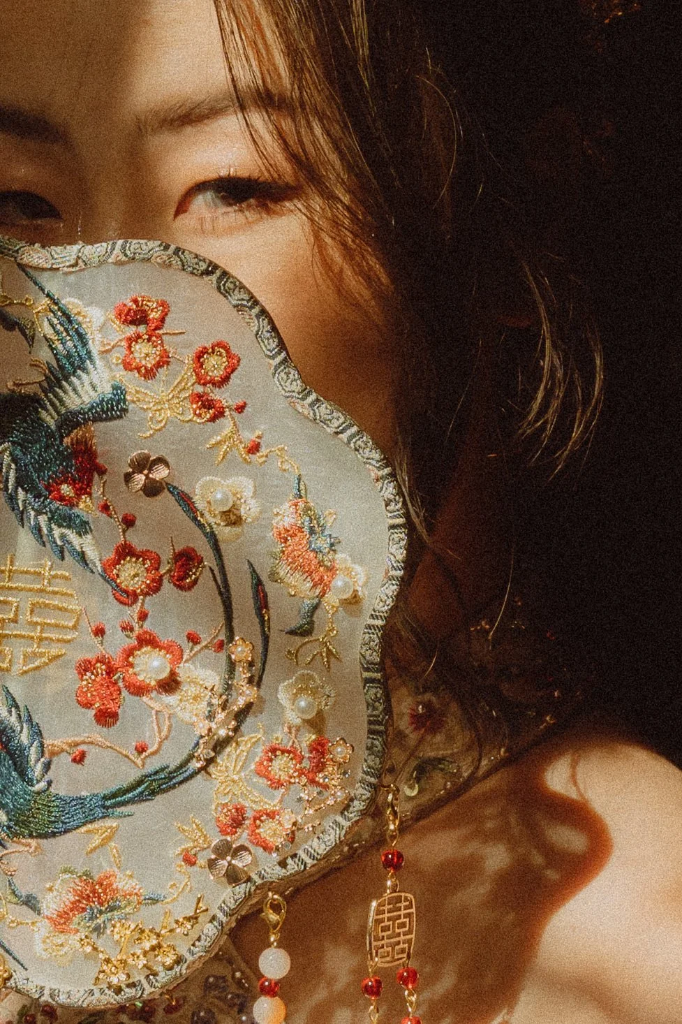 A woman holding an embroidered fan with floral and bird designs in front of her face, revealing her eye and part of her face. She wears jewelry with red beads.