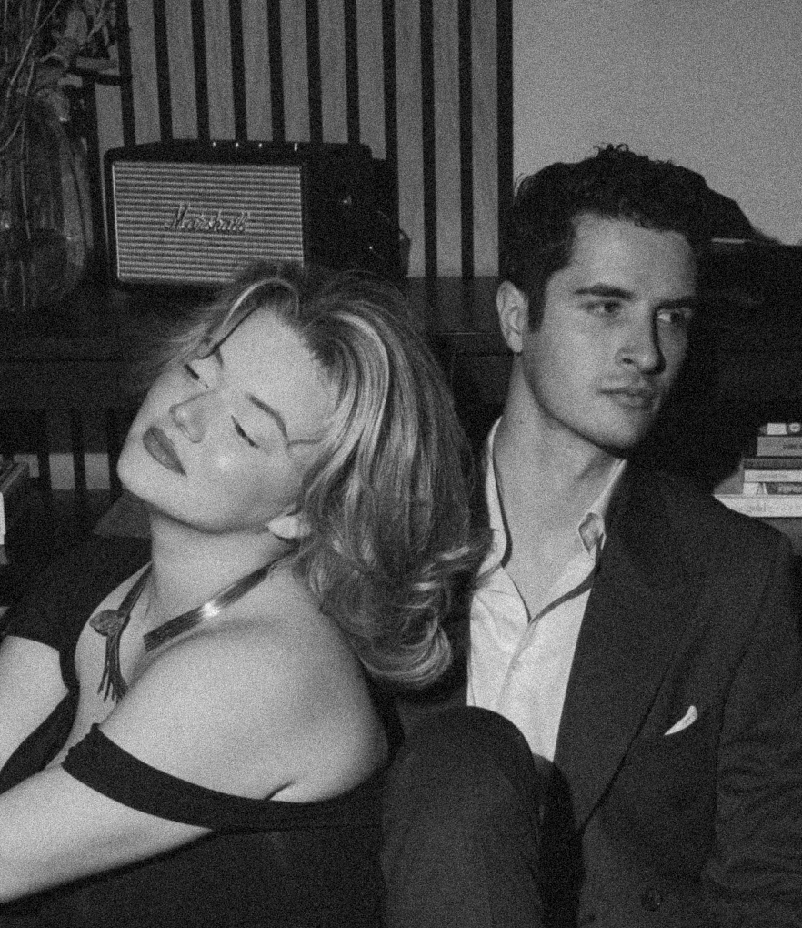 A black and white photo of a woman with short, wavy hair with her eyes closed and a man with short hair sitting beside her, both dressed in semi-formal attire, sitting indoors with a vintage radio and books in the background.