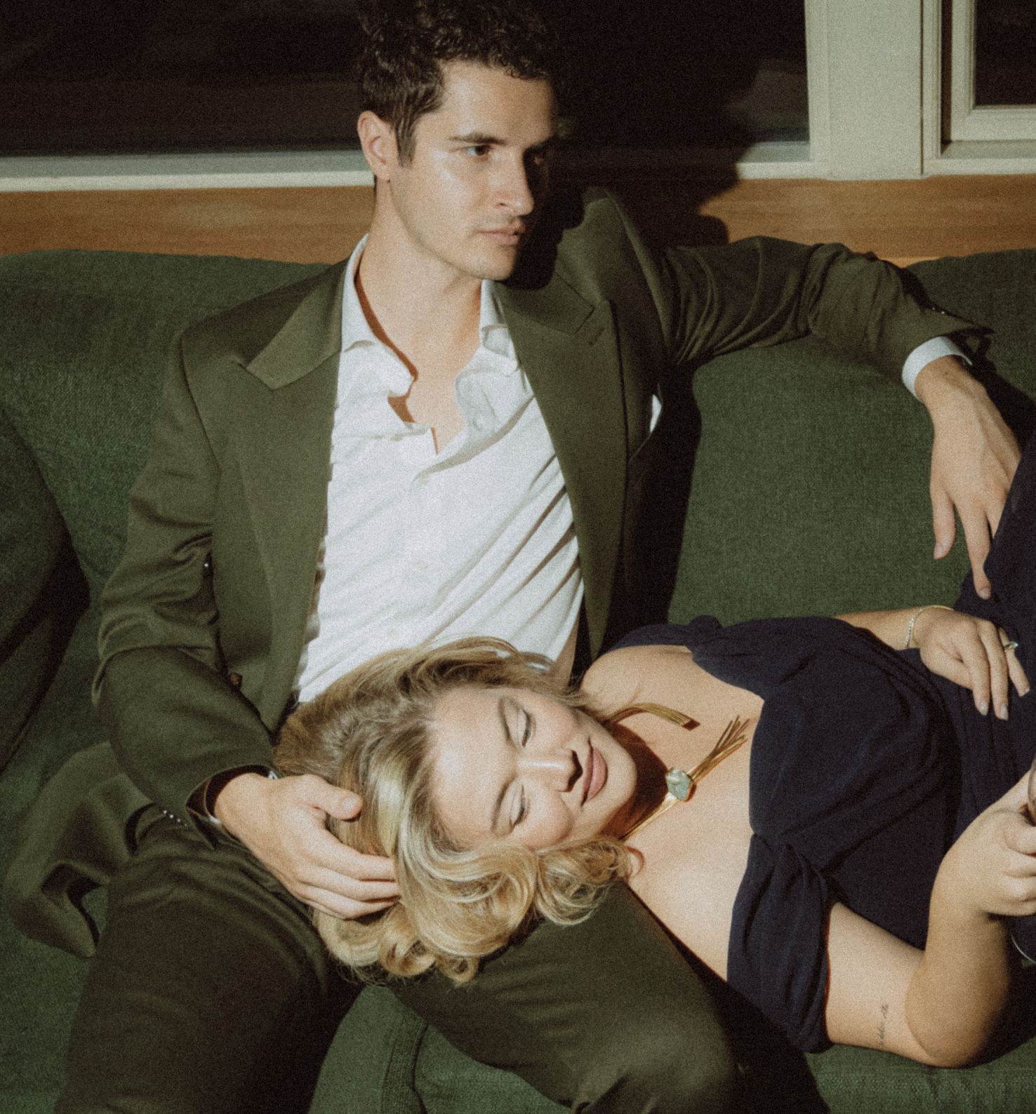 A young man in a suit is sitting on a green sofa with his eyes closed, appearing to be sleeping or resting. A young woman with blonde hair, also in dark clothing, is lying next to him with her eyes closed and head resting on his lap, suggesting a moment of relaxation or repose.