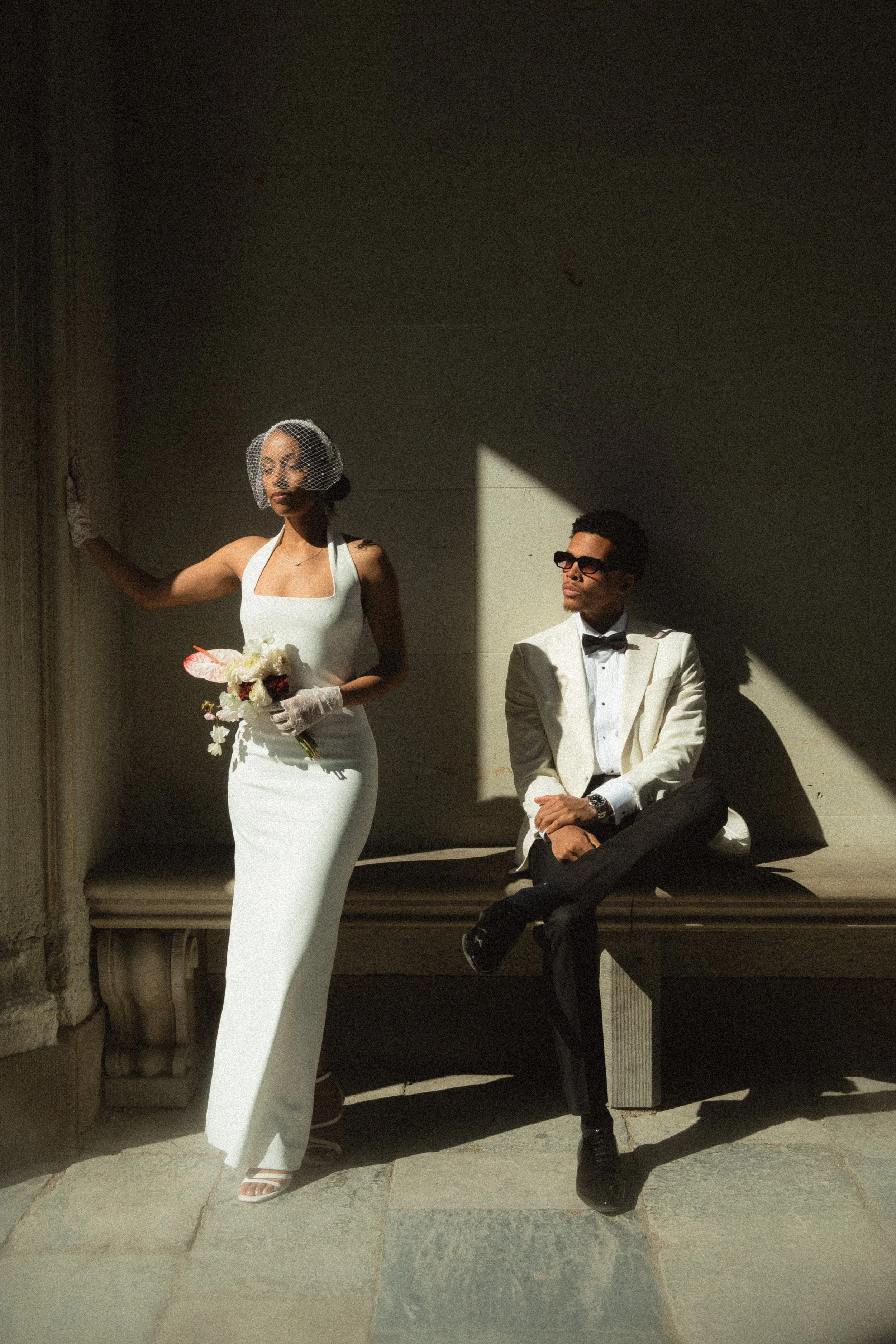 A woman dressed in a white wedding gown standing and holding a bouquet of flowers, wearing a birdcage veil. A man in a tuxedo with sunglasses is sitting on a bench beside her, with shadows cast on the wall behind them.