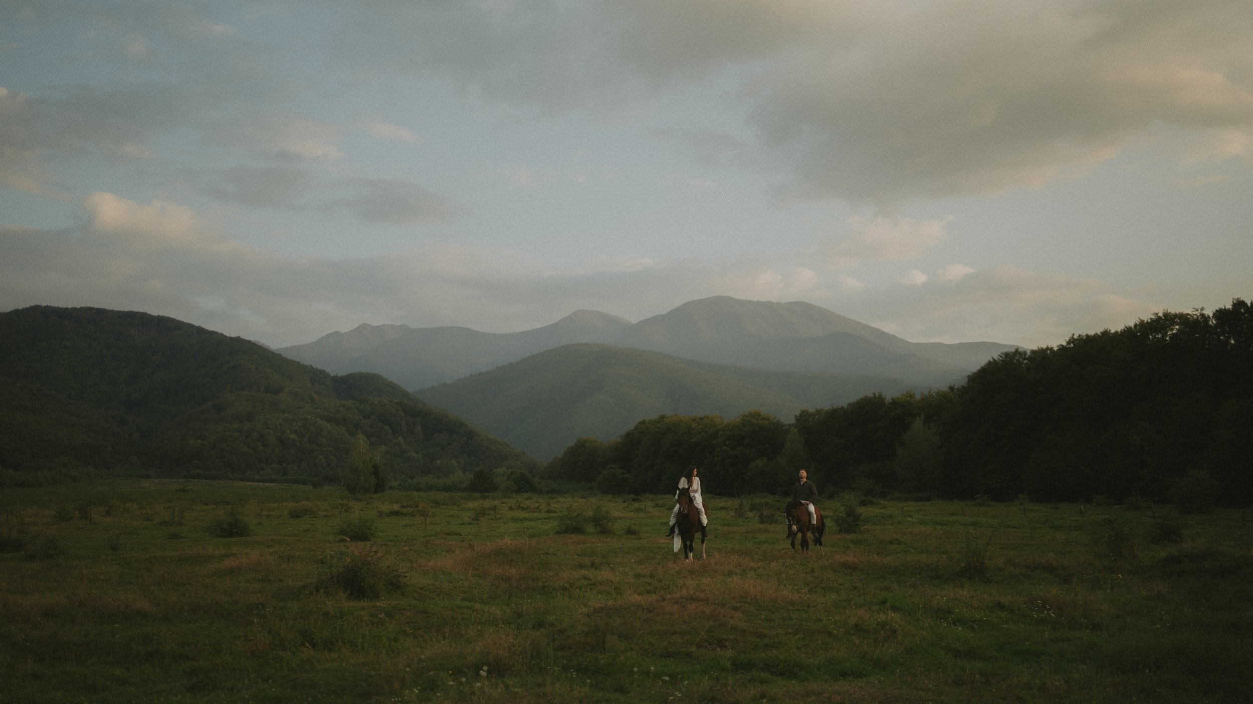Two people riding horses across a grassy field with mountain ranges and a partly cloudy sky in the background.