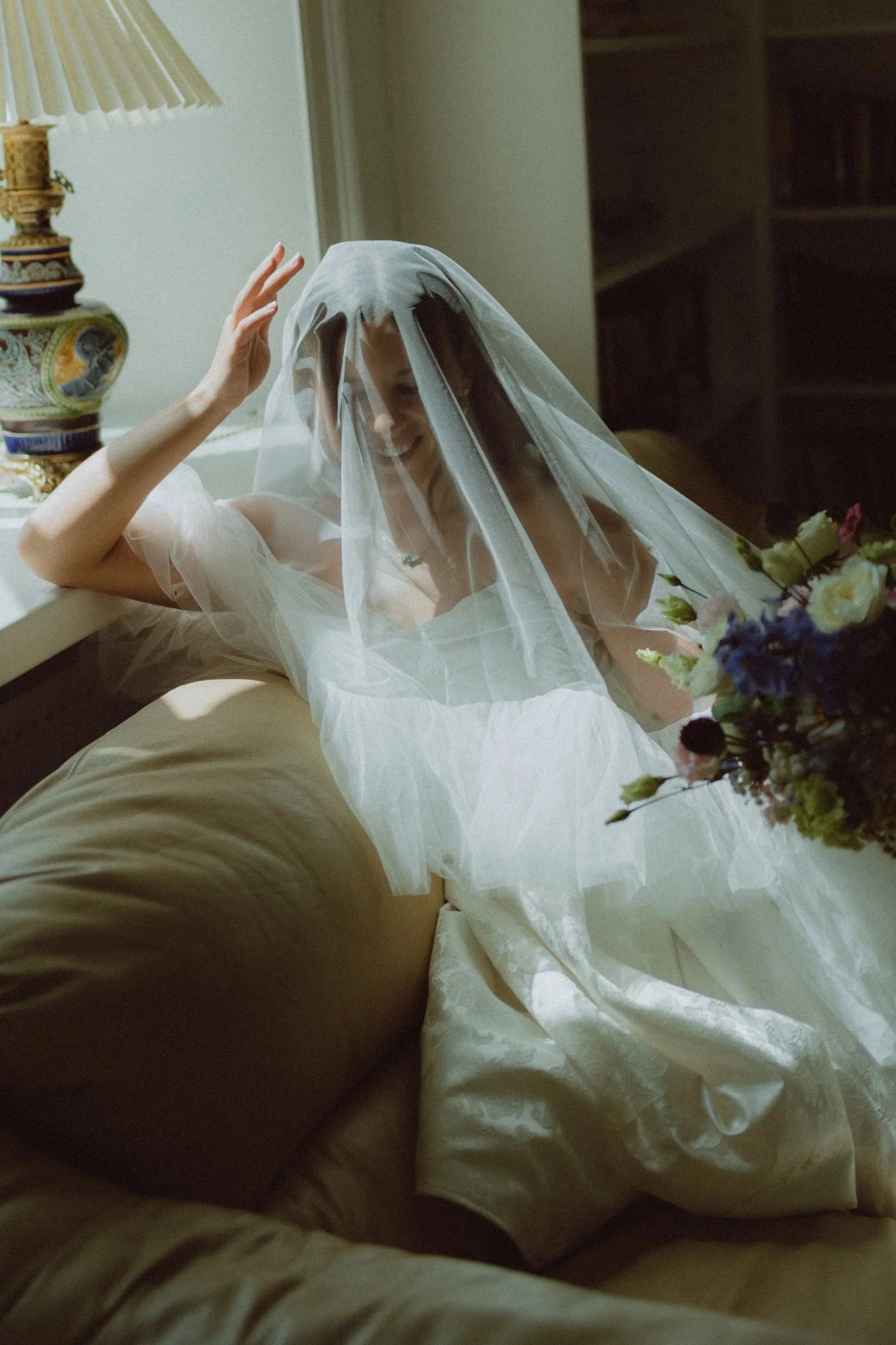 A woman with a veil on her head, sitting on a bed and smiling, surrounded by flowers, with a lamp and bookshelf in the background.