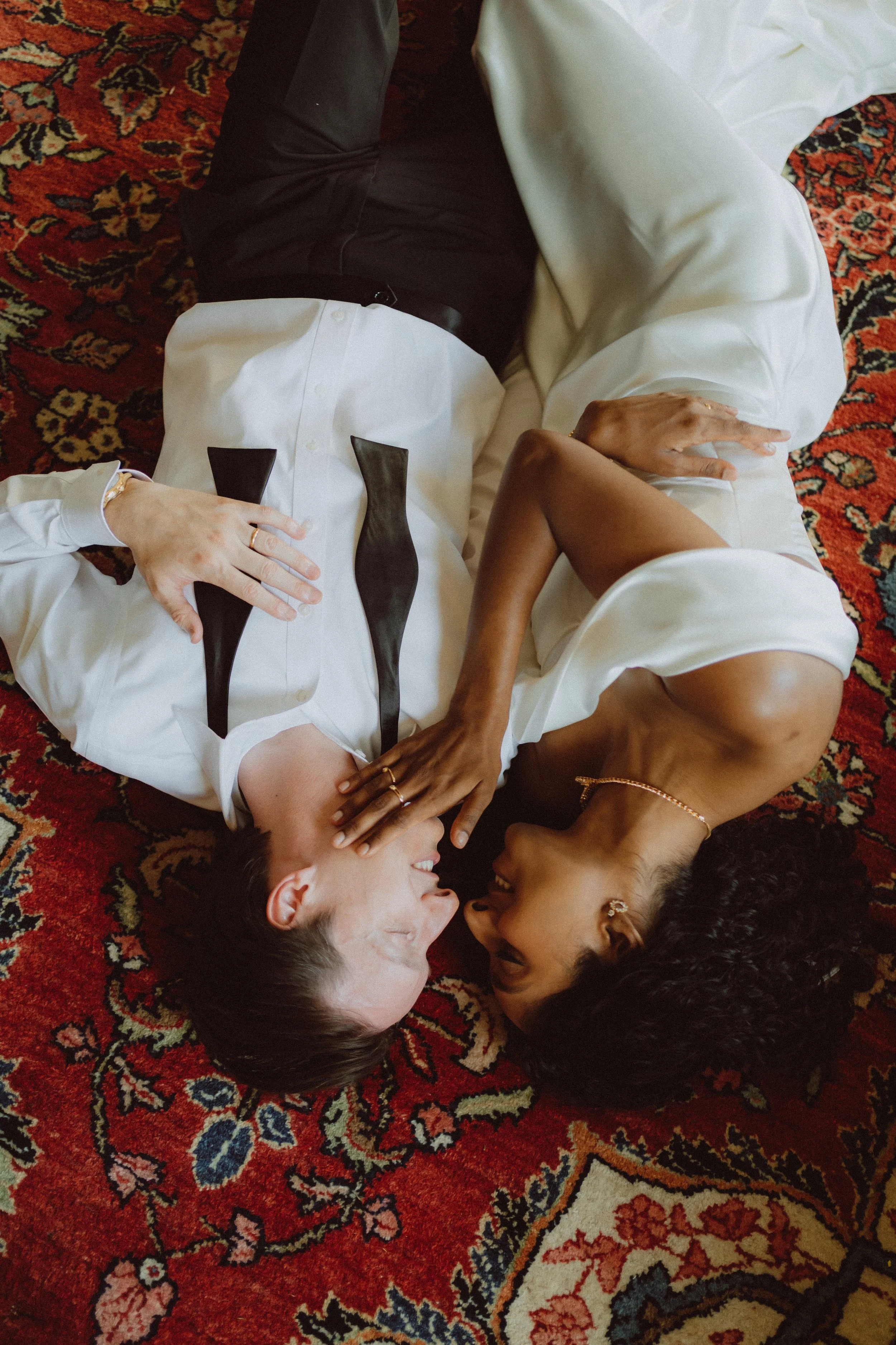 Two women and a man lying on a colorful patterned carpet, facing each other, smiling, with one woman wearing a white shirt with a black tie design and the other woman in a white dress. They appear to be happy and engaging in a moment of connection.
