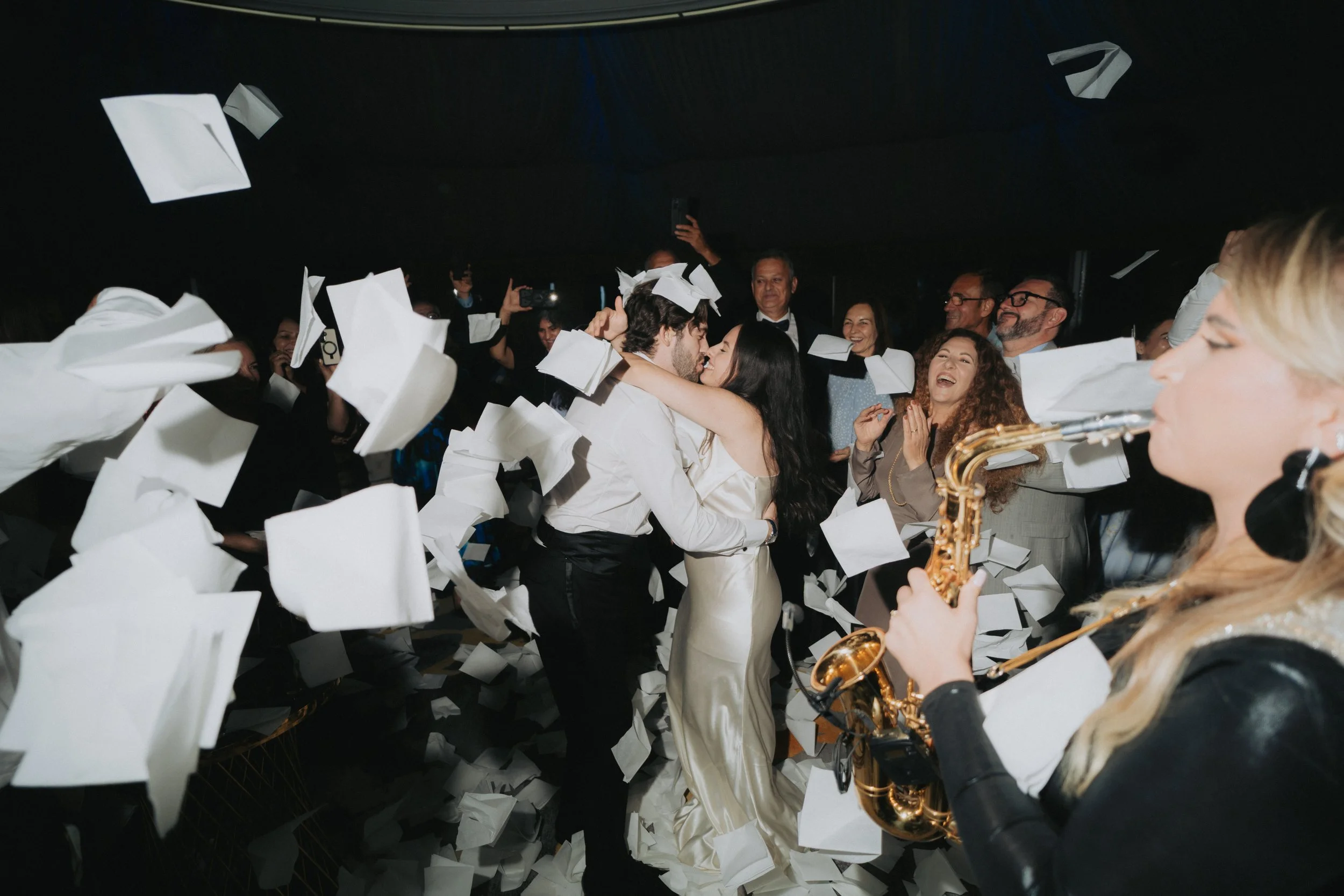 Couple kissing amidst falling papers at a celebration, with guests smiling and a saxophonist playing in the foreground.