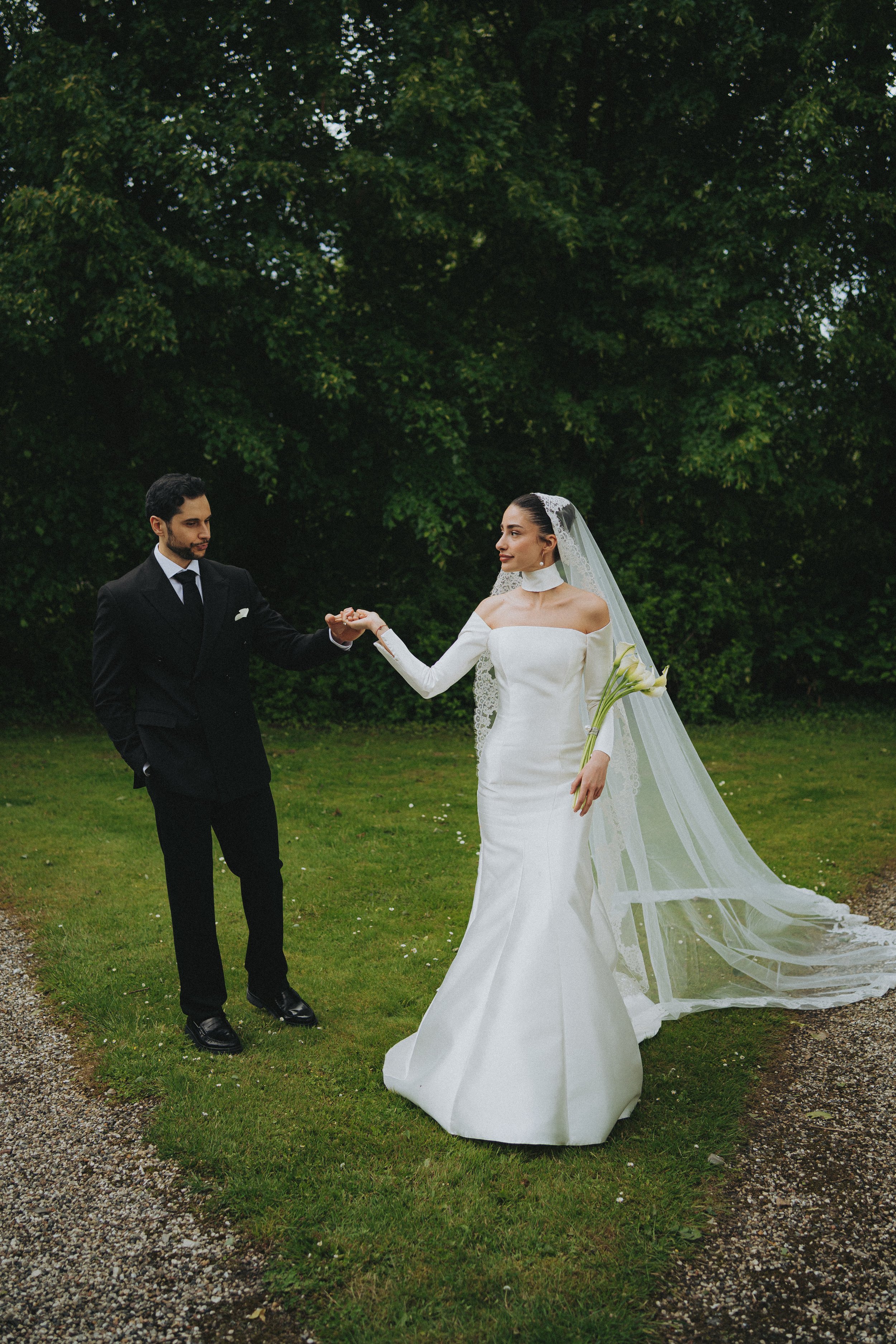 A bride in a long white wedding dress holding a bouquet of calla lilies and a groom in a black suit, exchanging rings outdoors in front of green trees.