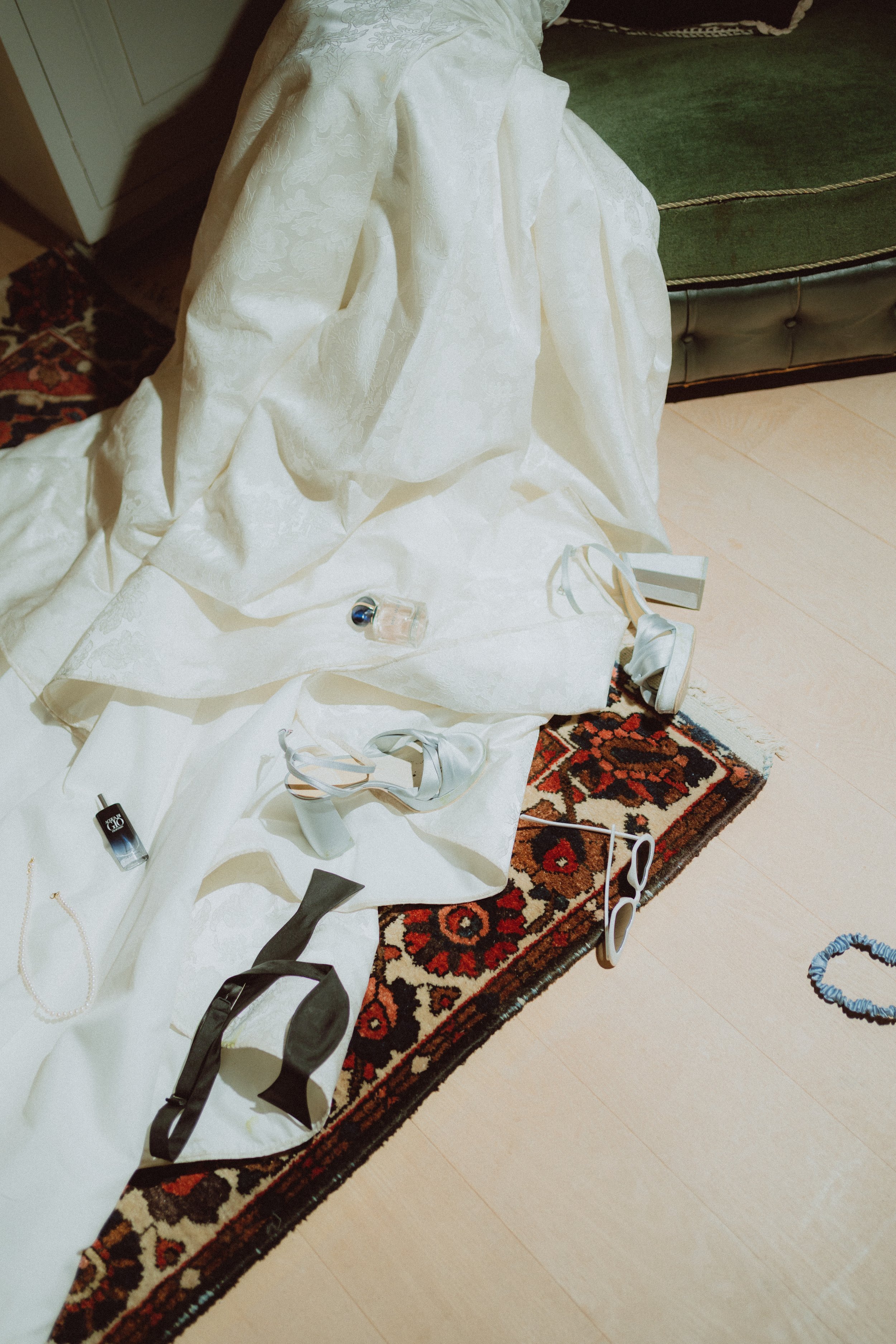 A patterned rug with a white fabric covering a surface, scattered with shoes, jewelry, nail polish, and sunglasses, next to a green upholstered piece of furniture and beige tiled flooring.