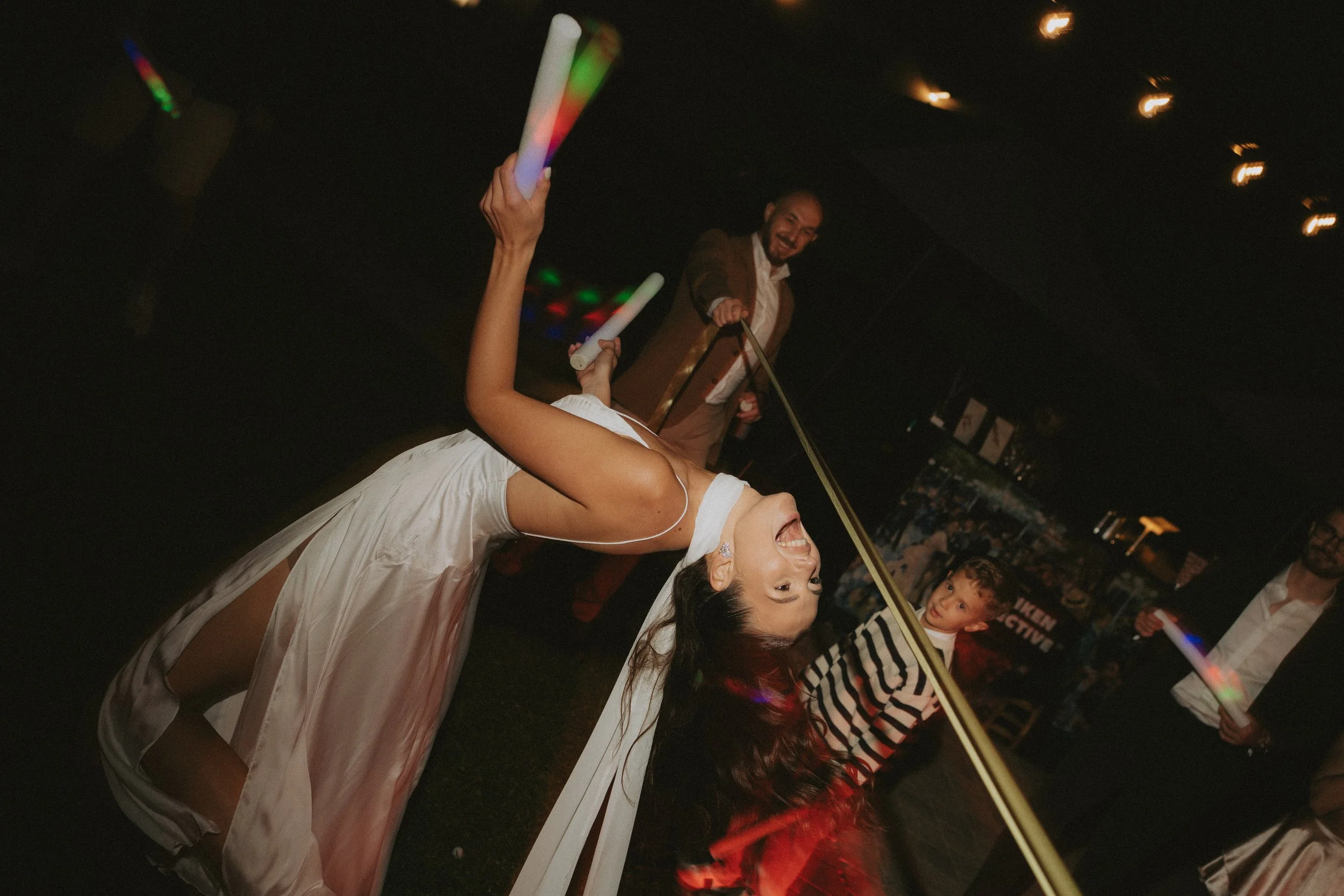 People celebrating at a party with glow sticks, including a woman in a satin dress and a man in a brown jacket, in a dimly lit room with string lights.