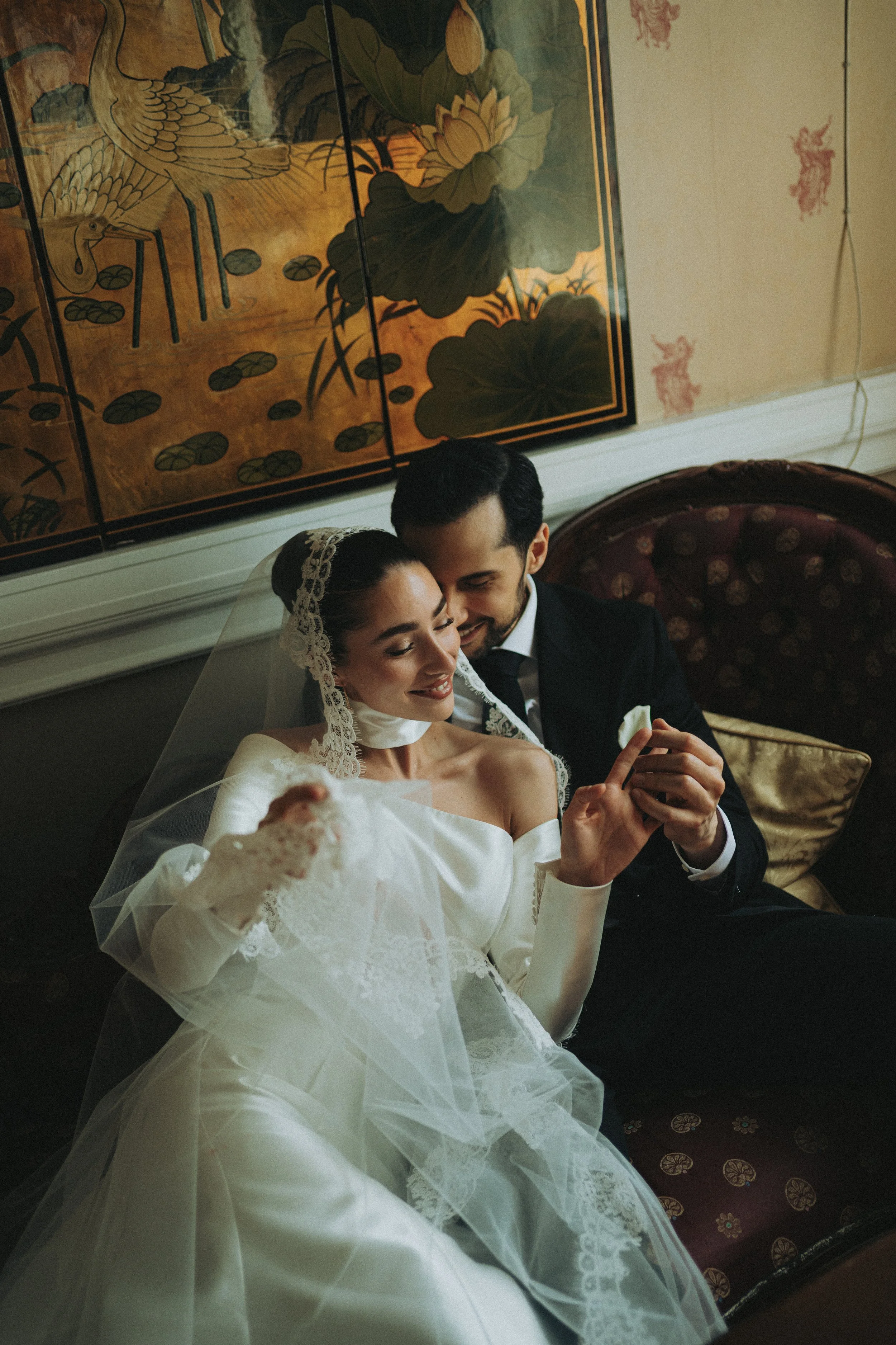 A bride and groom sitting together, smiling, with the bride in a white wedding dress and lace gloves, and the groom in a black suit. They are holding hands and appear happy. The background features traditional Asian-style artwork with birds and lotus