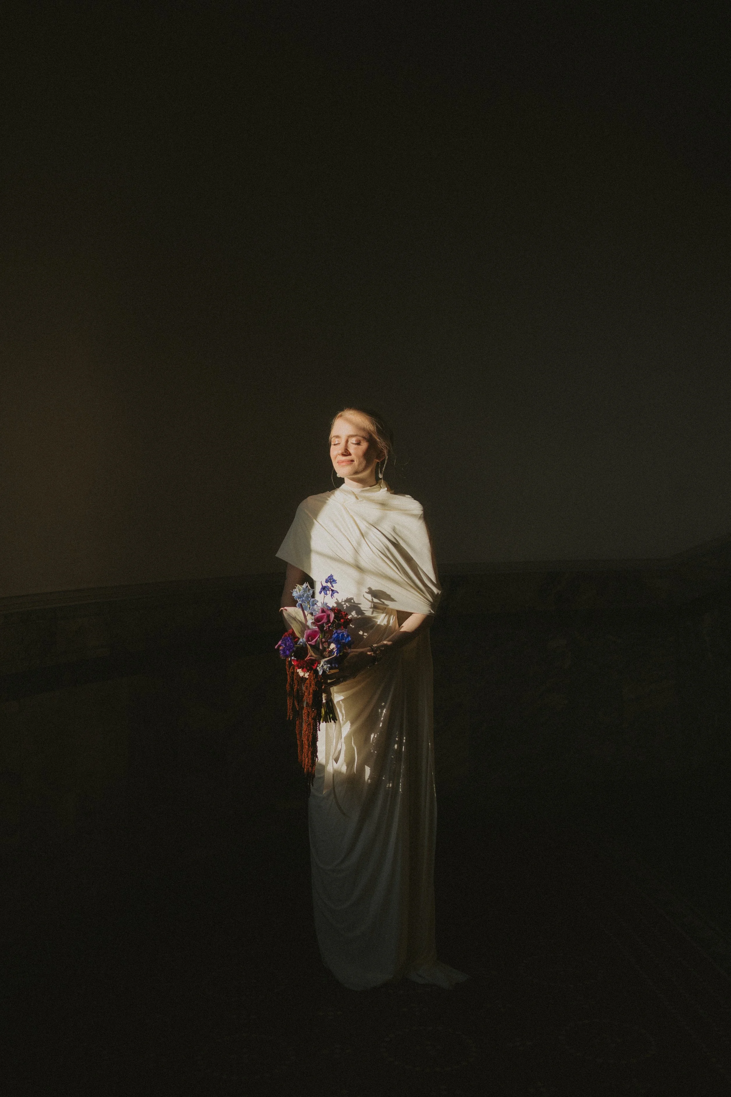 A woman in a flowing, elegant beige dress holds a bouquet of colorful flowers and stands in a dark environment with soft lighting highlighting her face and upper body.