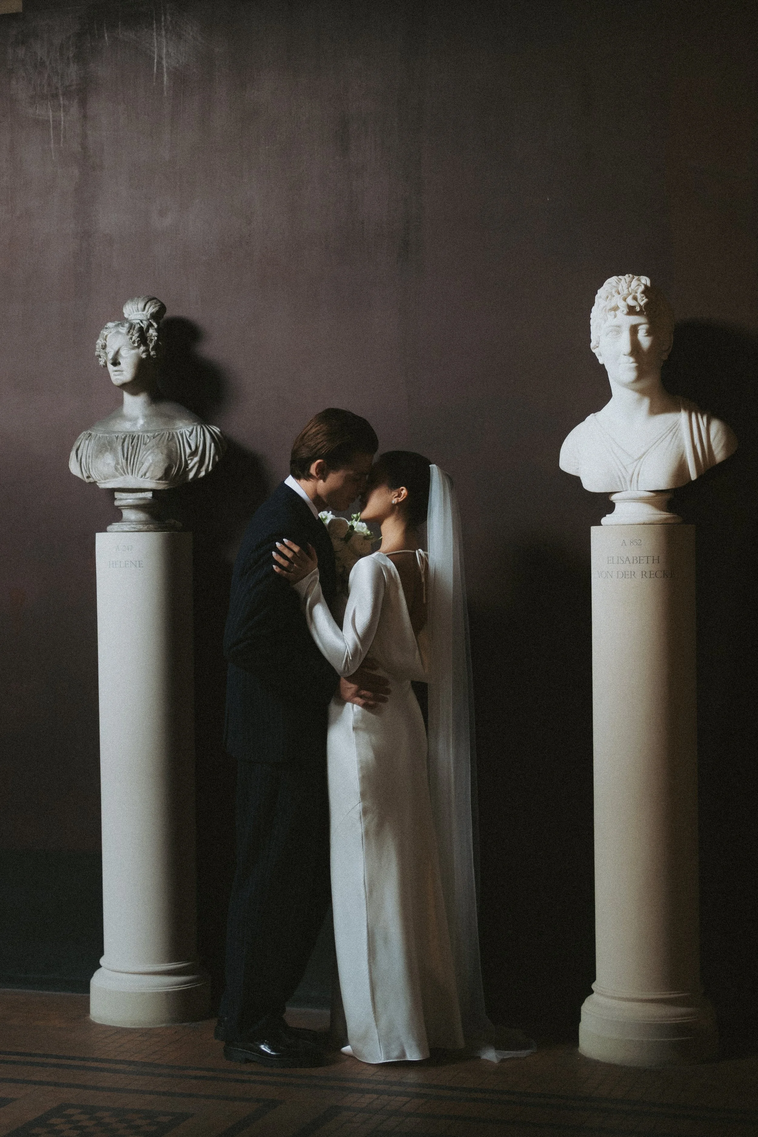 A bride and groom standing close together, holding hands and leaning on each other inside a museum with classical bust sculptures on white pedestals. The groom is dressed in a dark suit, and the bride is wearing a white dress and veil. The sculptures