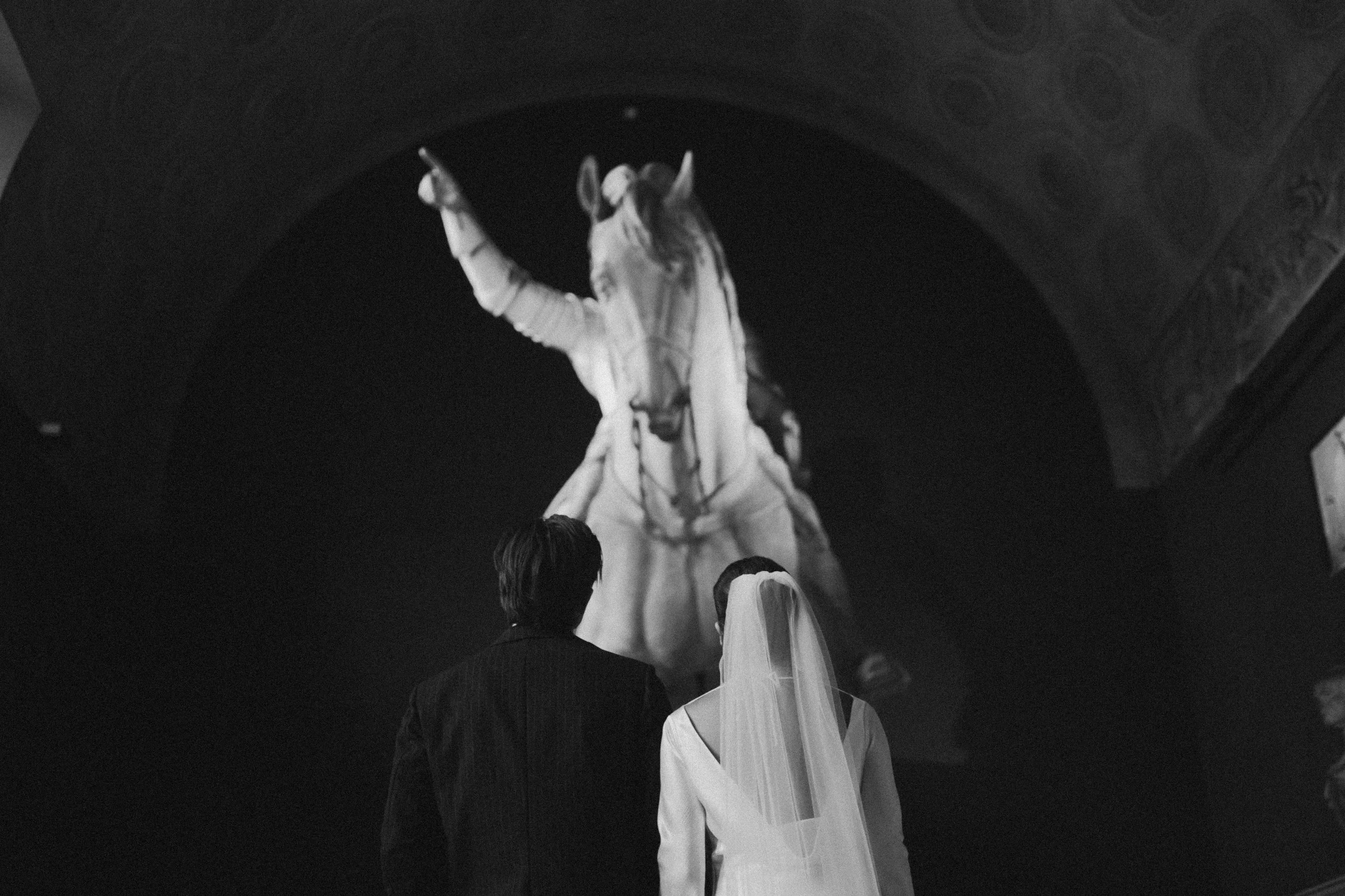 Black and white photograph of a sculpture of a person on horseback, with audience members in the foreground, set in an interior with an ornate ceiling.