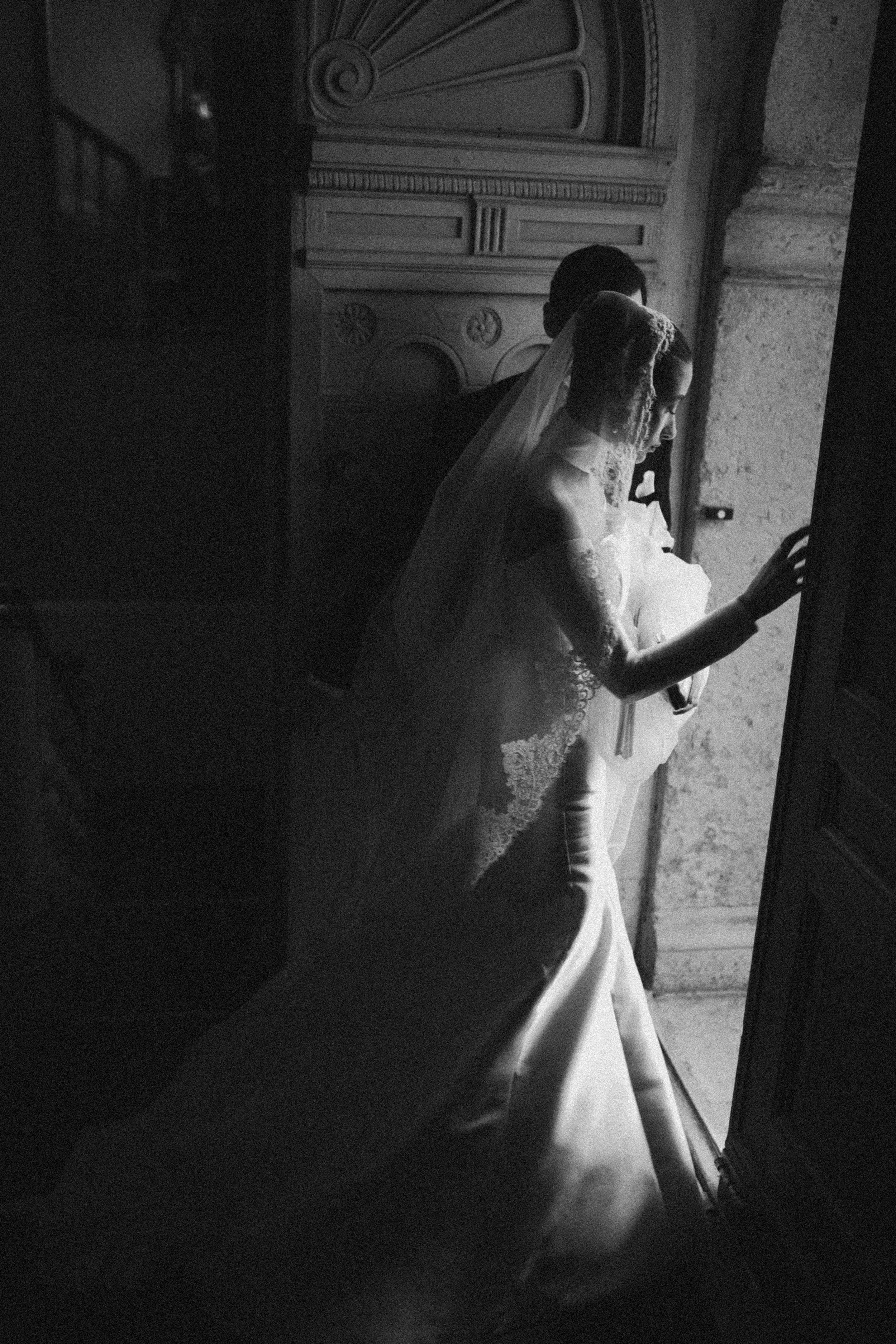 Black and white photo of a woman in a wedding dress with lace details, wearing a veil, looking out a window with a thoughtful expression, standing beside a man whose face is not visible.