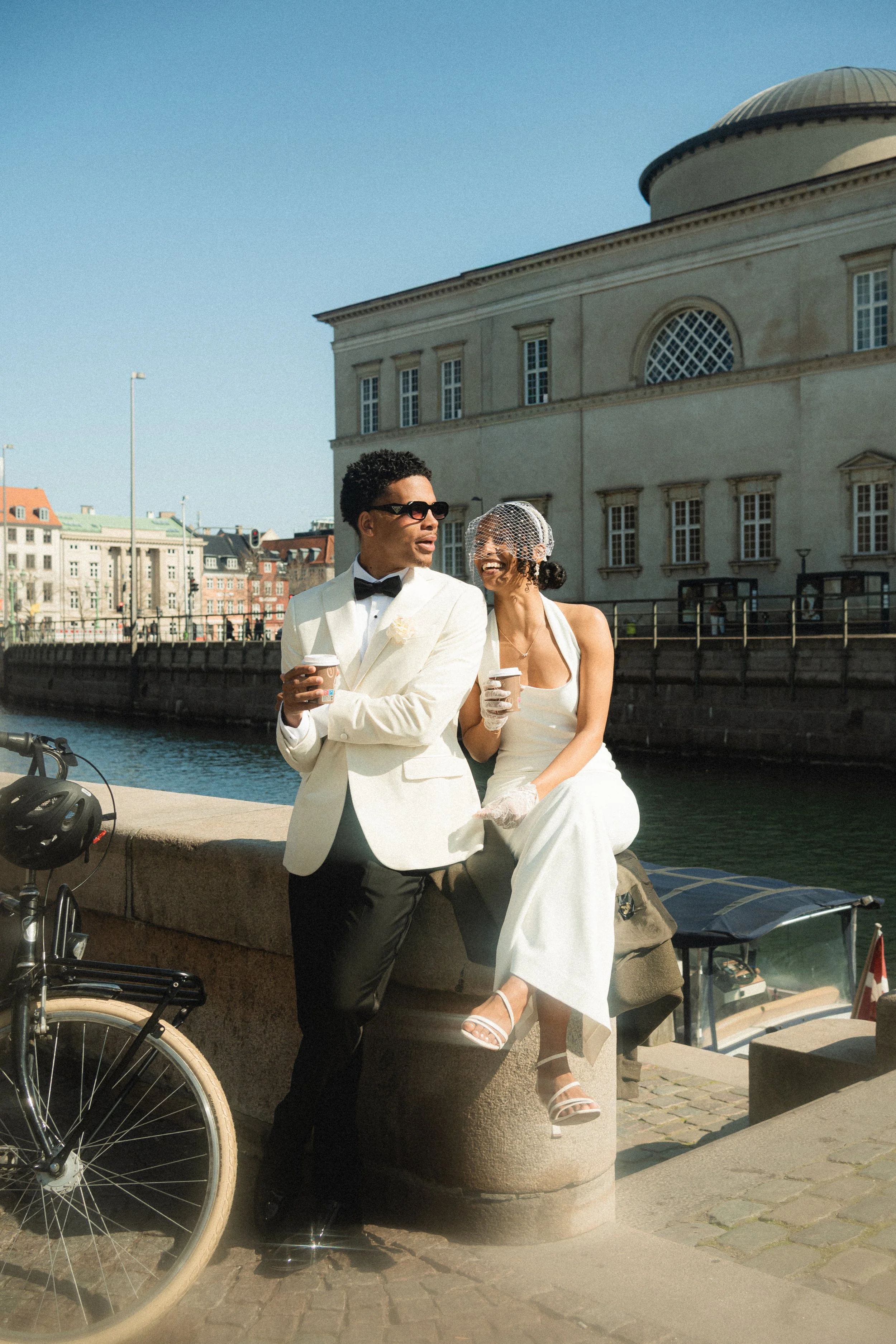 A man and a woman dressed in formal wedding attire sitting on a stone ledge by a canal, with buildings and a church in the background. The man is wearing a white tuxedo with a bow tie, and the woman is wearing a white wedding dress with a netted veil