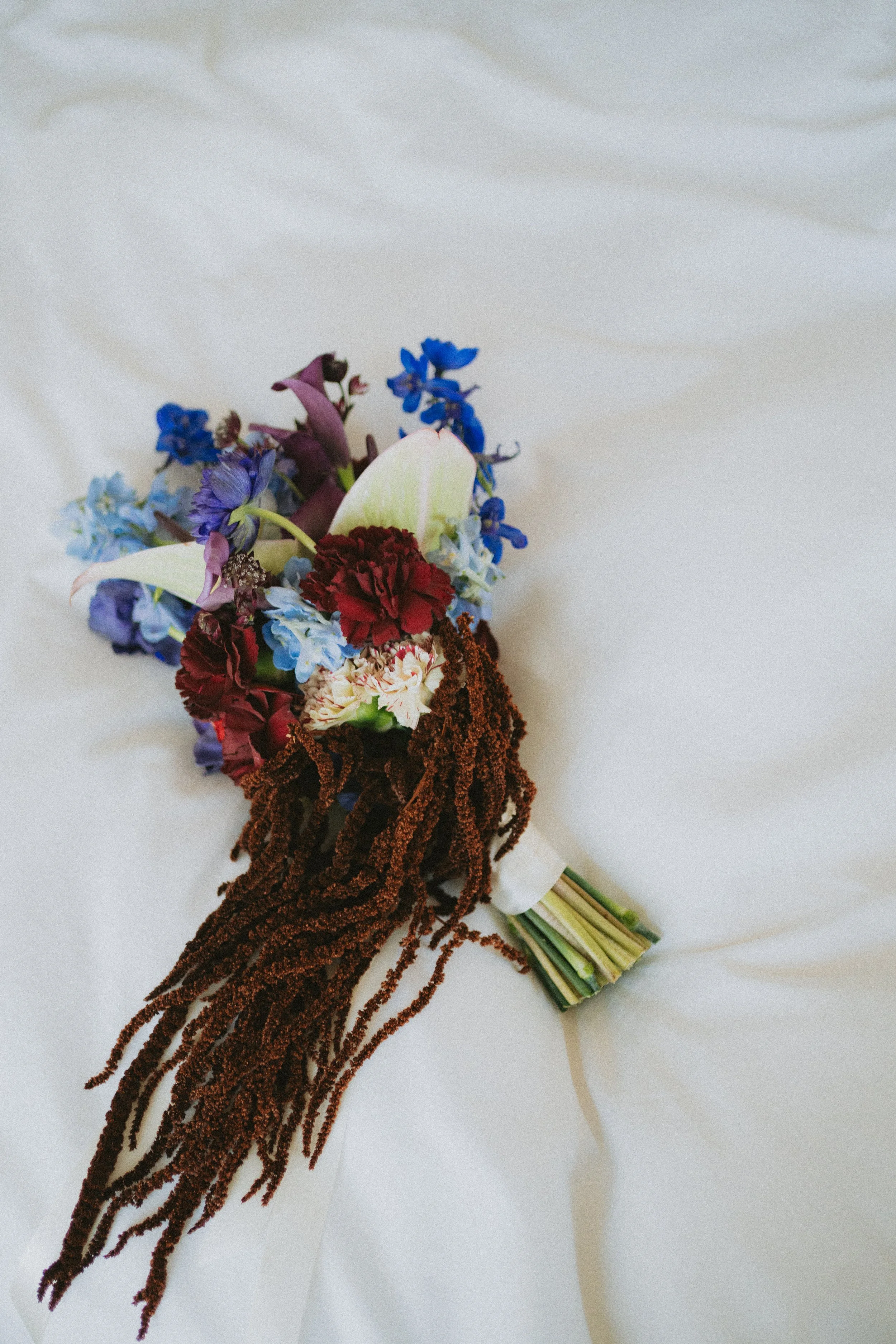 A floral bouquet on a white surface, consisting of blue, purple, red, and white flowers with green stems, wrapped with what appears to be a striped ribbon.