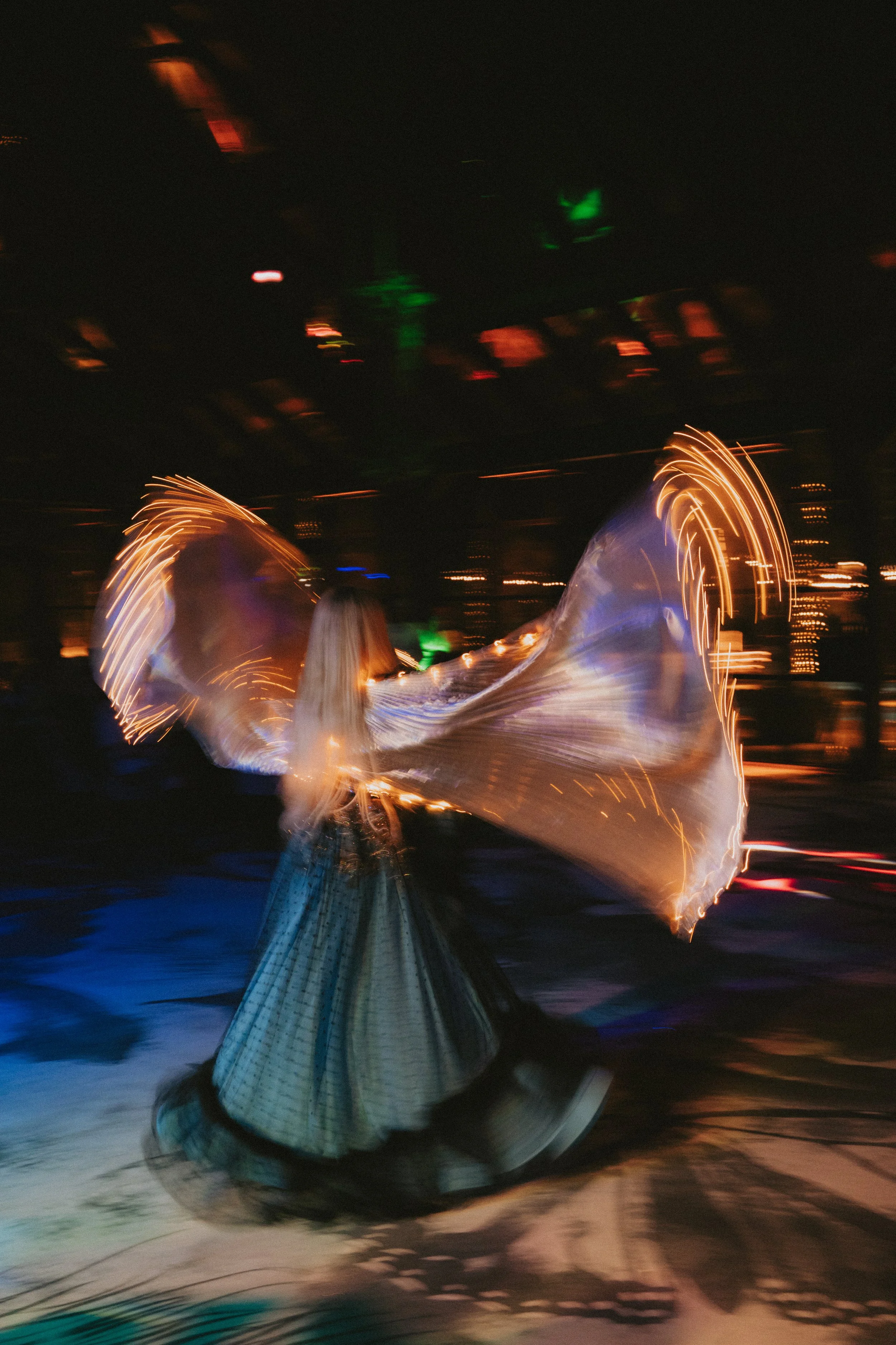 A person is spinning a piece of fabric that is illuminated, creating a light trail effect against a dark background, with city lights and green and red lights visible in the distance.