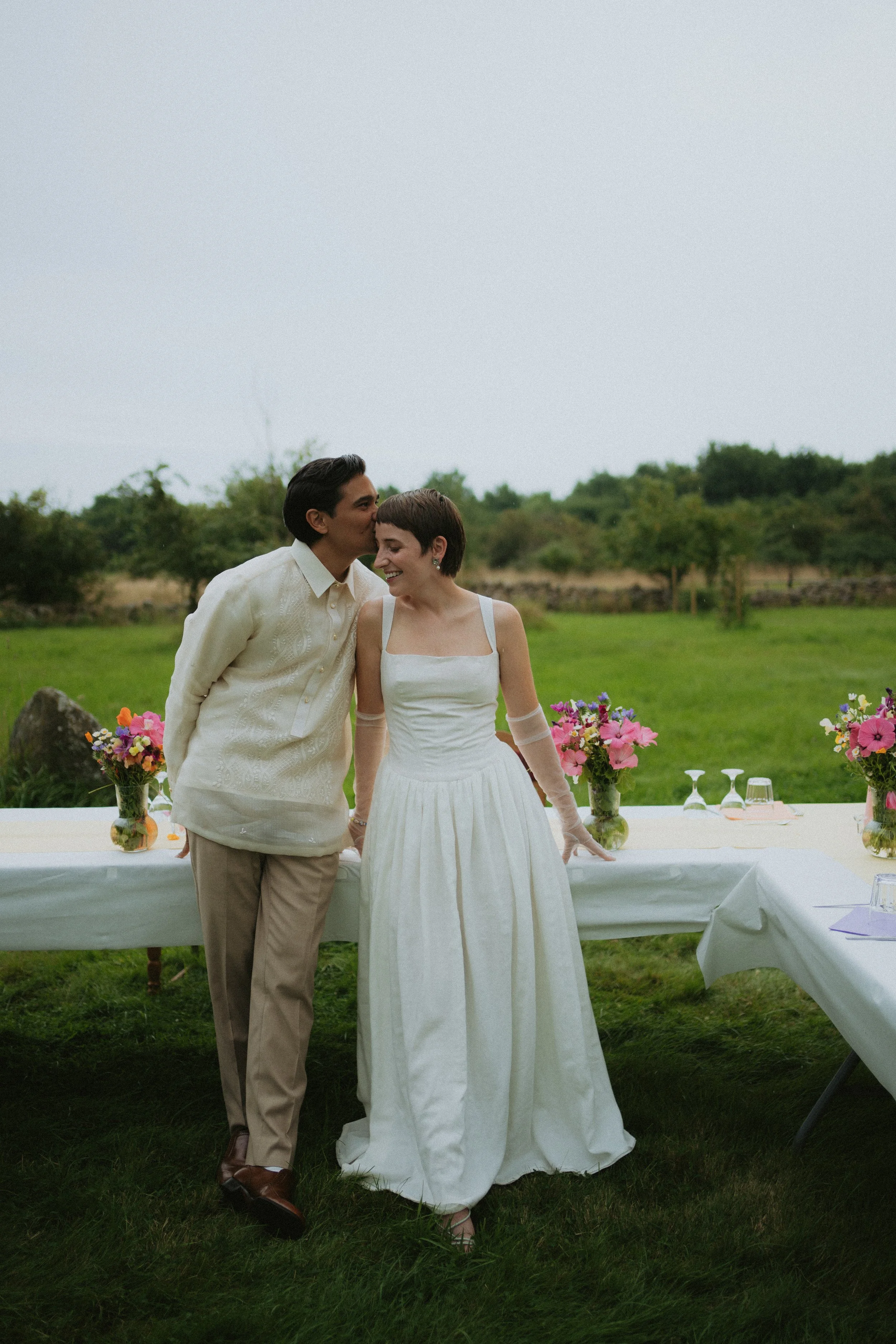 A couple dressed in wedding attire standing outdoors at a wedding reception, with a table and colorful flower arrangements visible behind them.