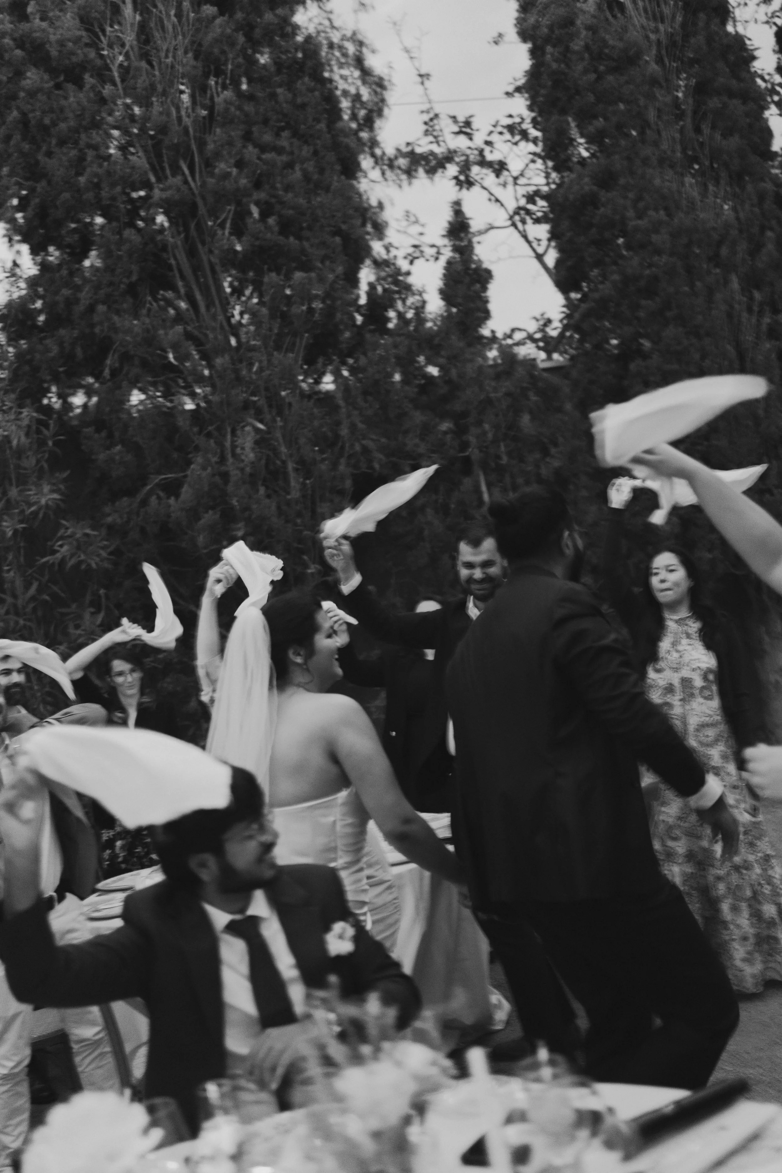 People dancing and socializing outdoors, with some wearing formal attire and others dressed casually, under a tree canopy.