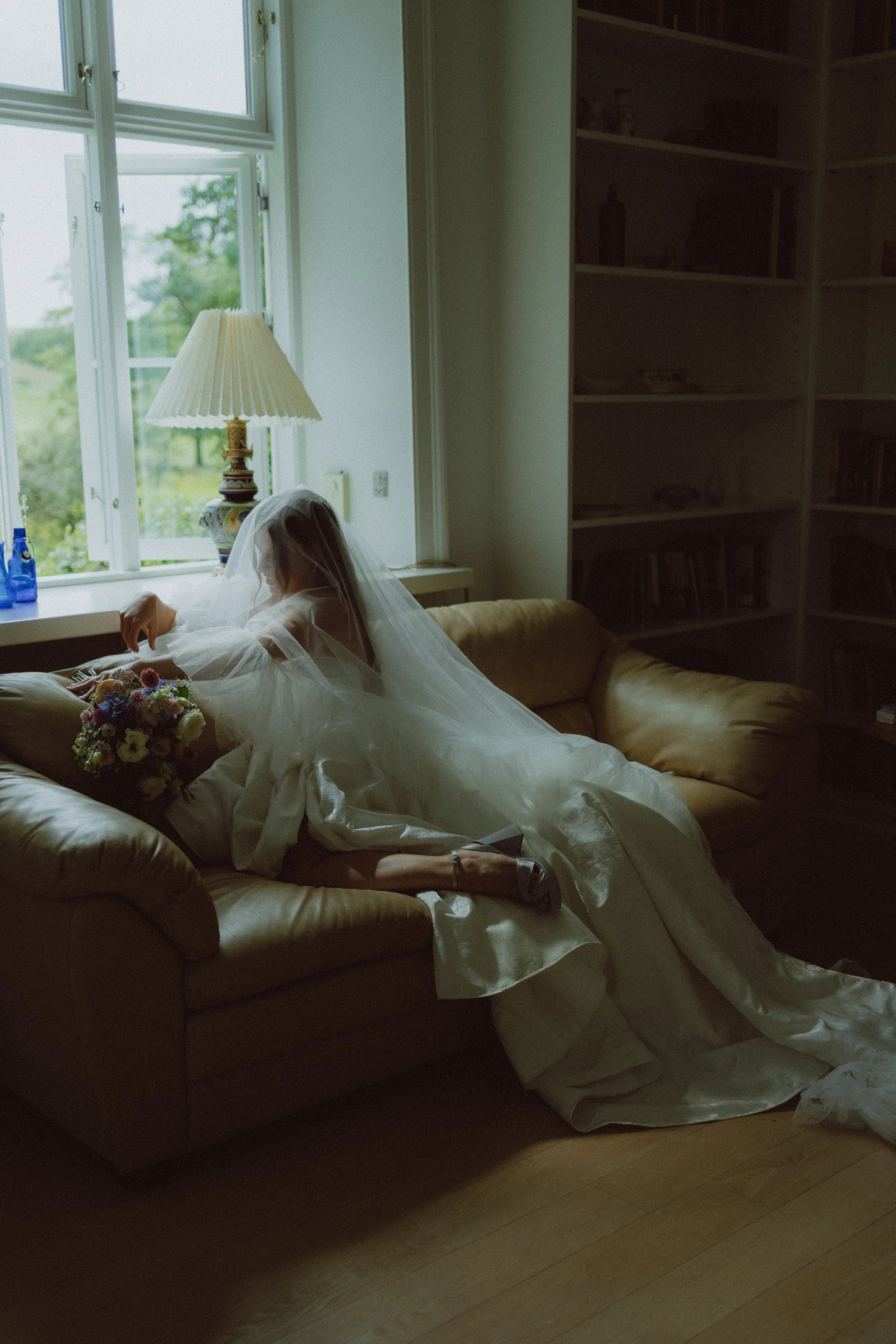 A woman in a wedding dress and veil is lying on a sofa, holding a bouquet of flowers, near a window with greenery outside, with a dark bookshelf behind her.