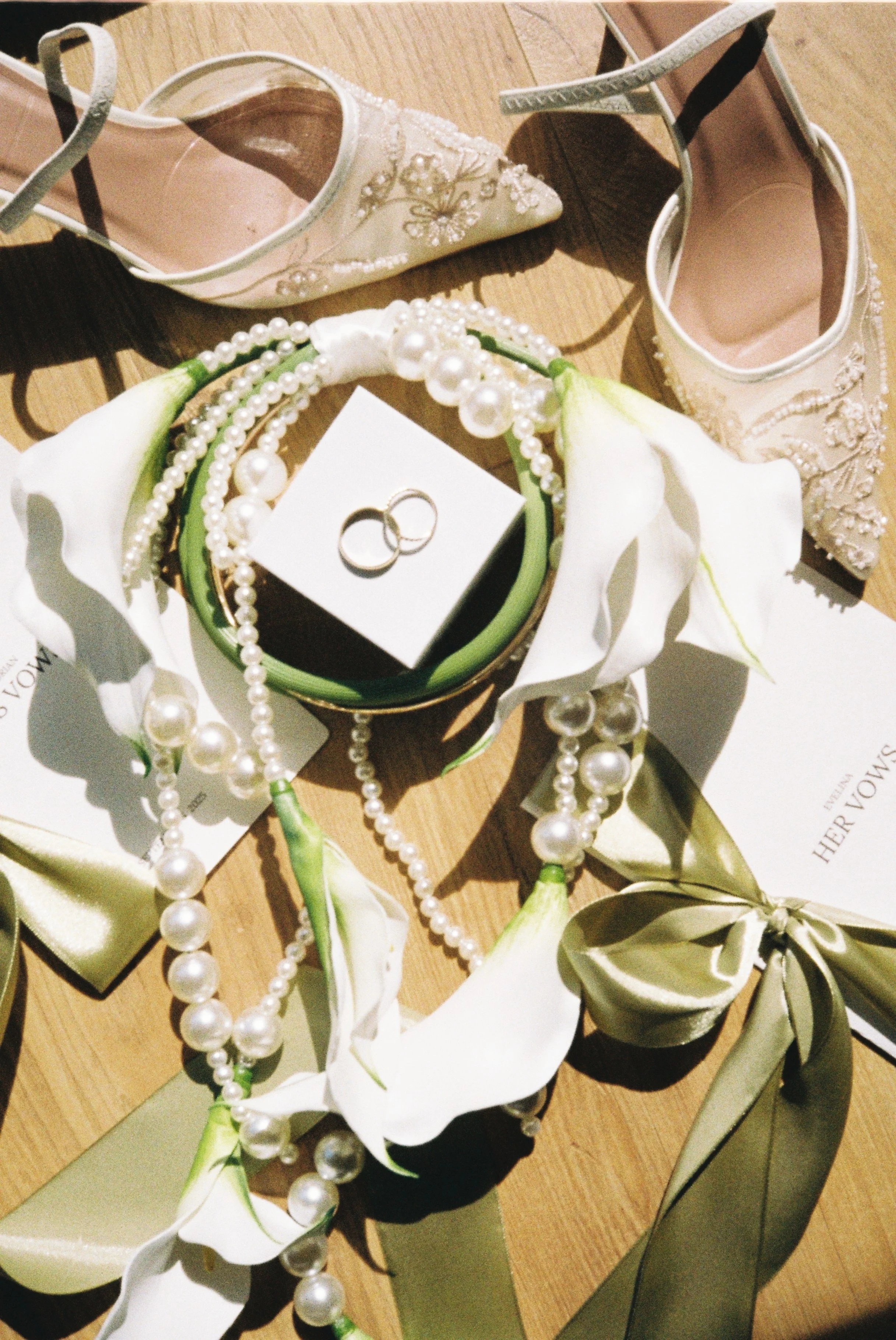 A pair of elegant beige high-heeled shoes with intricate beadwork, placed on a wooden surface. A string of pearls and a white calla lily flower are also in the scene, along with a pair of wedding rings on a small black box.