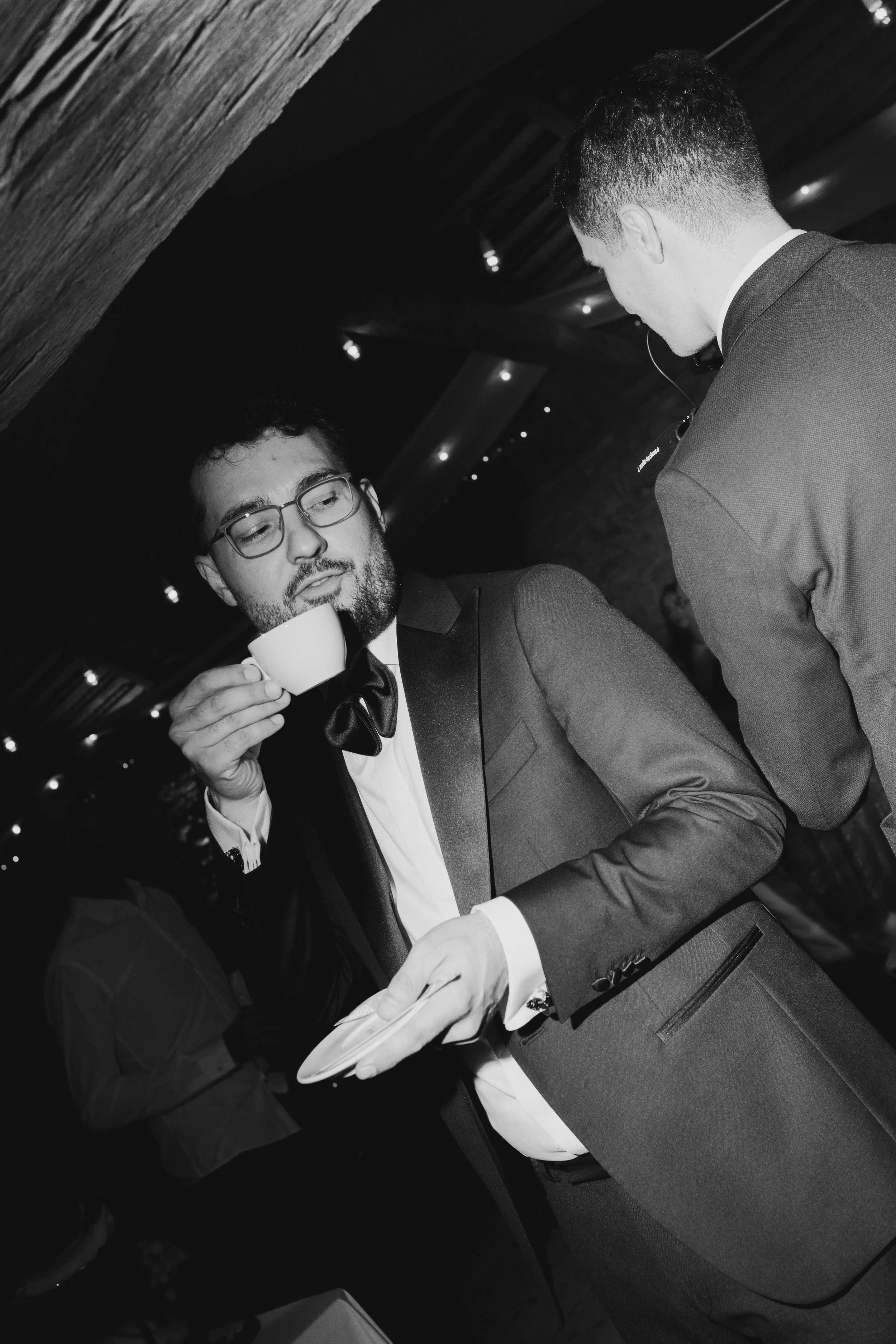 Two men in formal attire during a night event, one holding a cup and the other holding a plate, engaging in conversation inside a dimly lit venue with wooden ceiling beams.