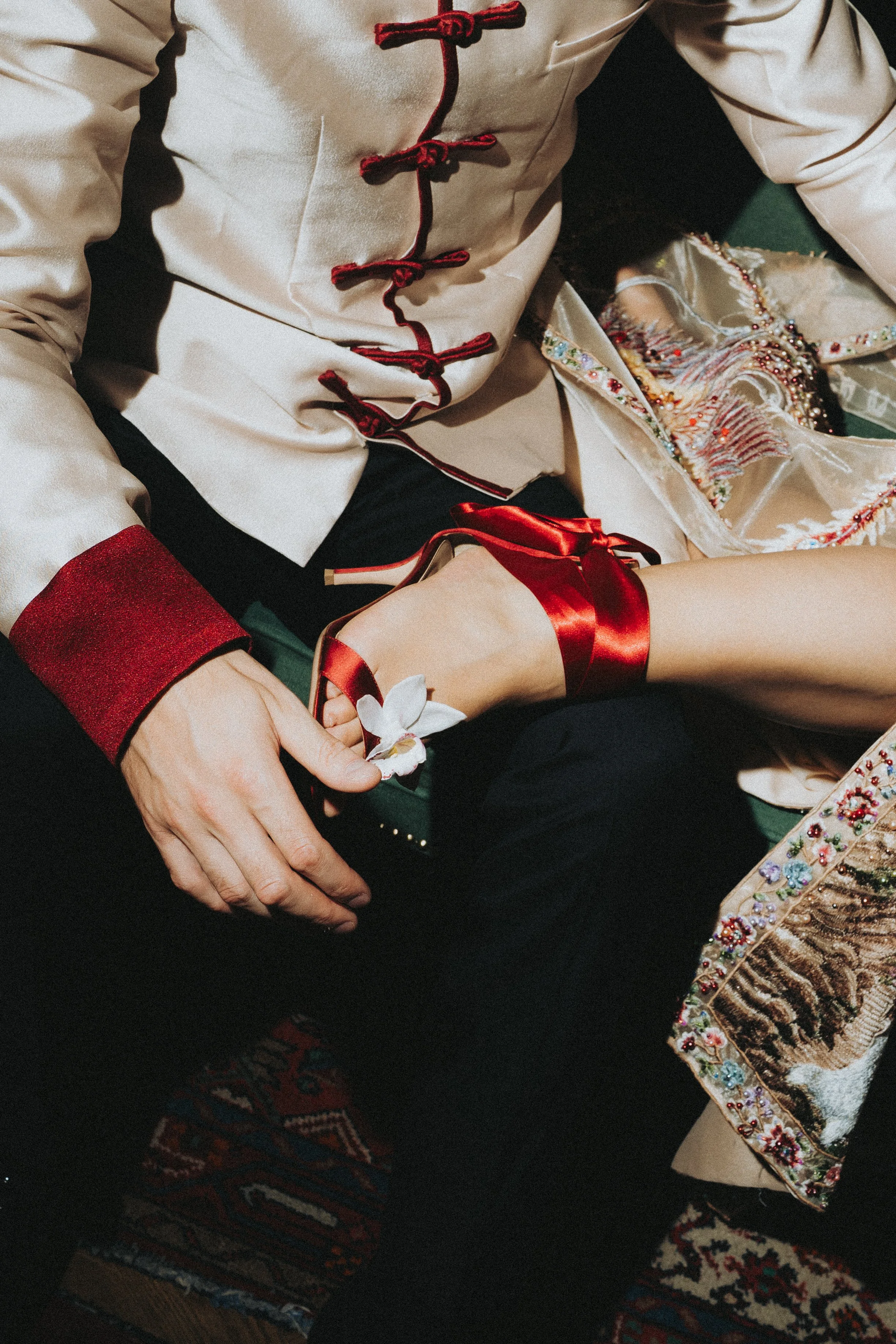 Close-up of a person wearing traditional Asian clothing with intricate embroidery, beadwork, and a red ribbon, relaxing on a patterned rug.