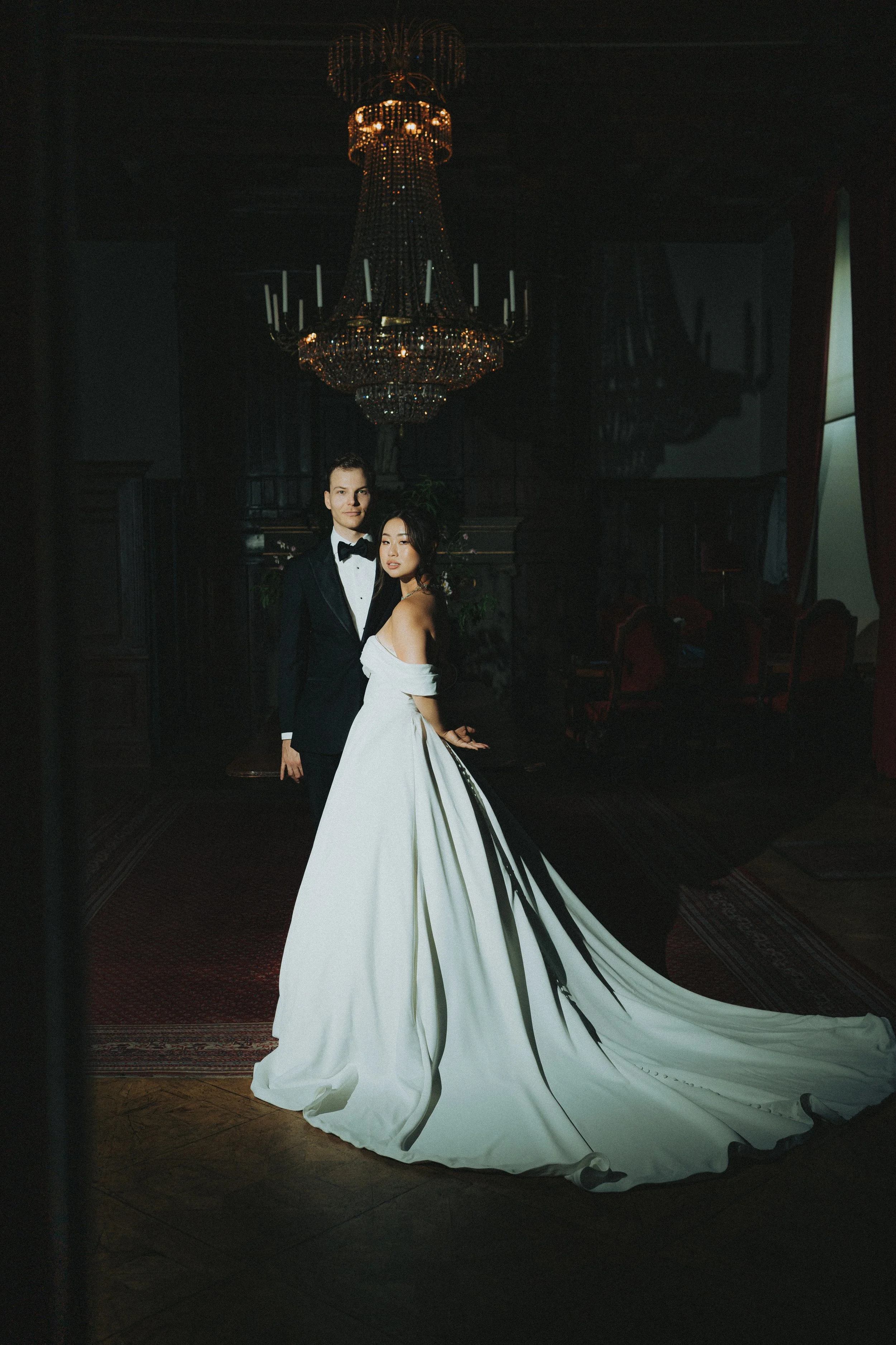 A man and woman dressed in formal attire standing together in a dimly lit room with a large chandelier overhead. The woman is wearing a white gown with a long train and the man is in a black tuxedo. The woman is slightly tilting her head and the man 