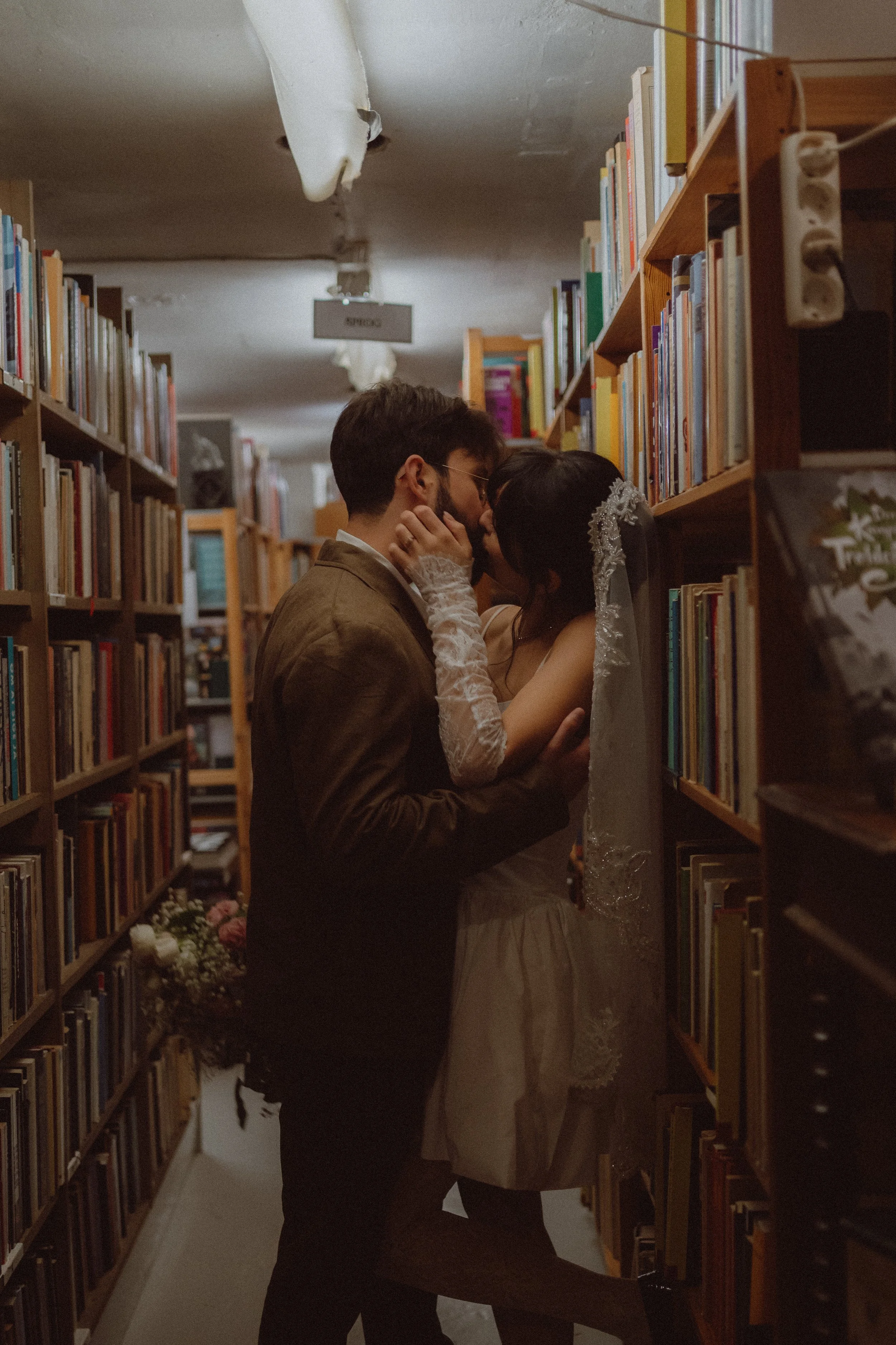 A man and woman, dressed in wedding attire, sharing an intimate moment in a library aisle with bookshelves on both sides.