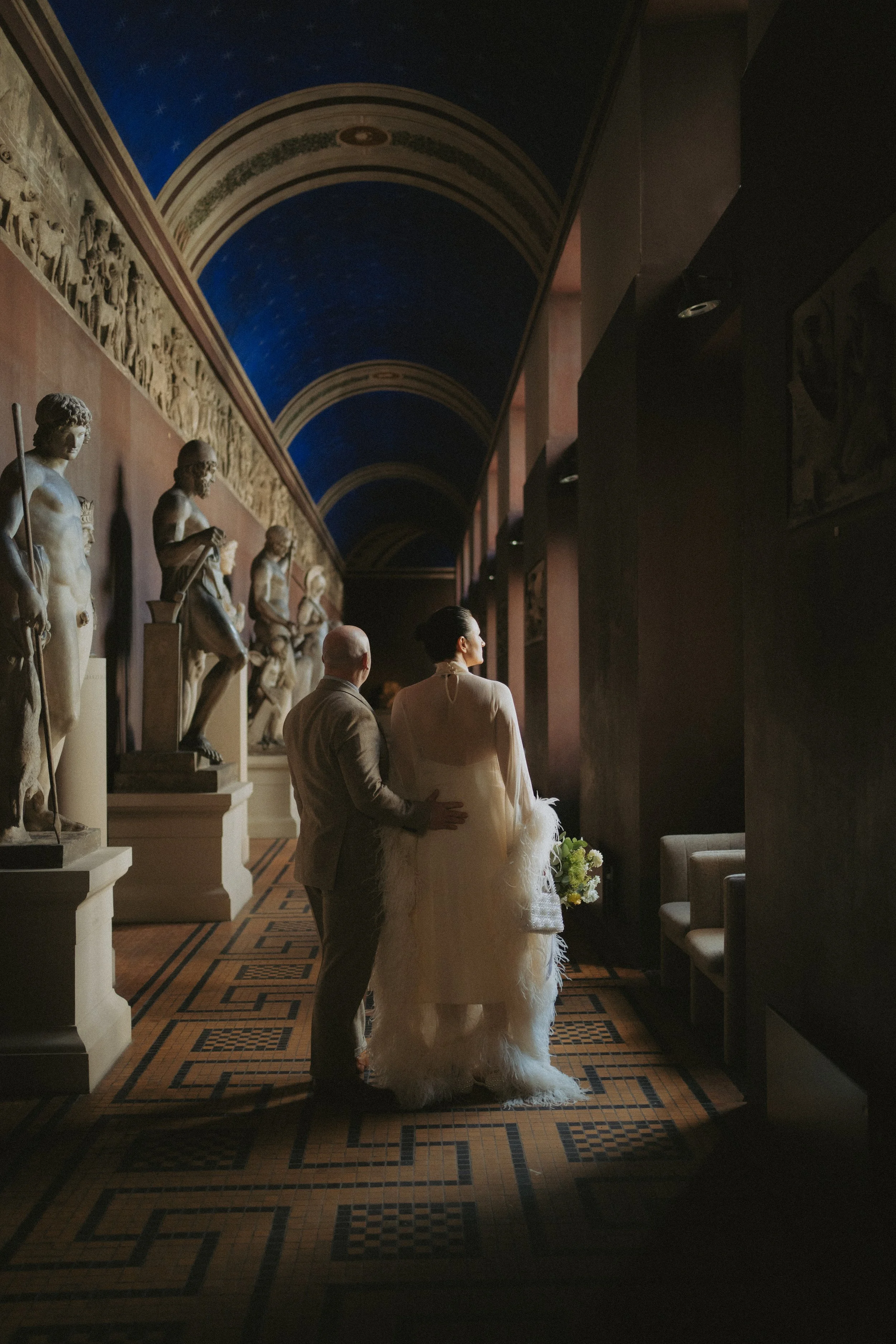 A bride and groom standing together in a hallway decorated with classical statues on the left and dark walls on the right. The hallway has a detailed tiled floor and a painted ceiling with arches and stars.