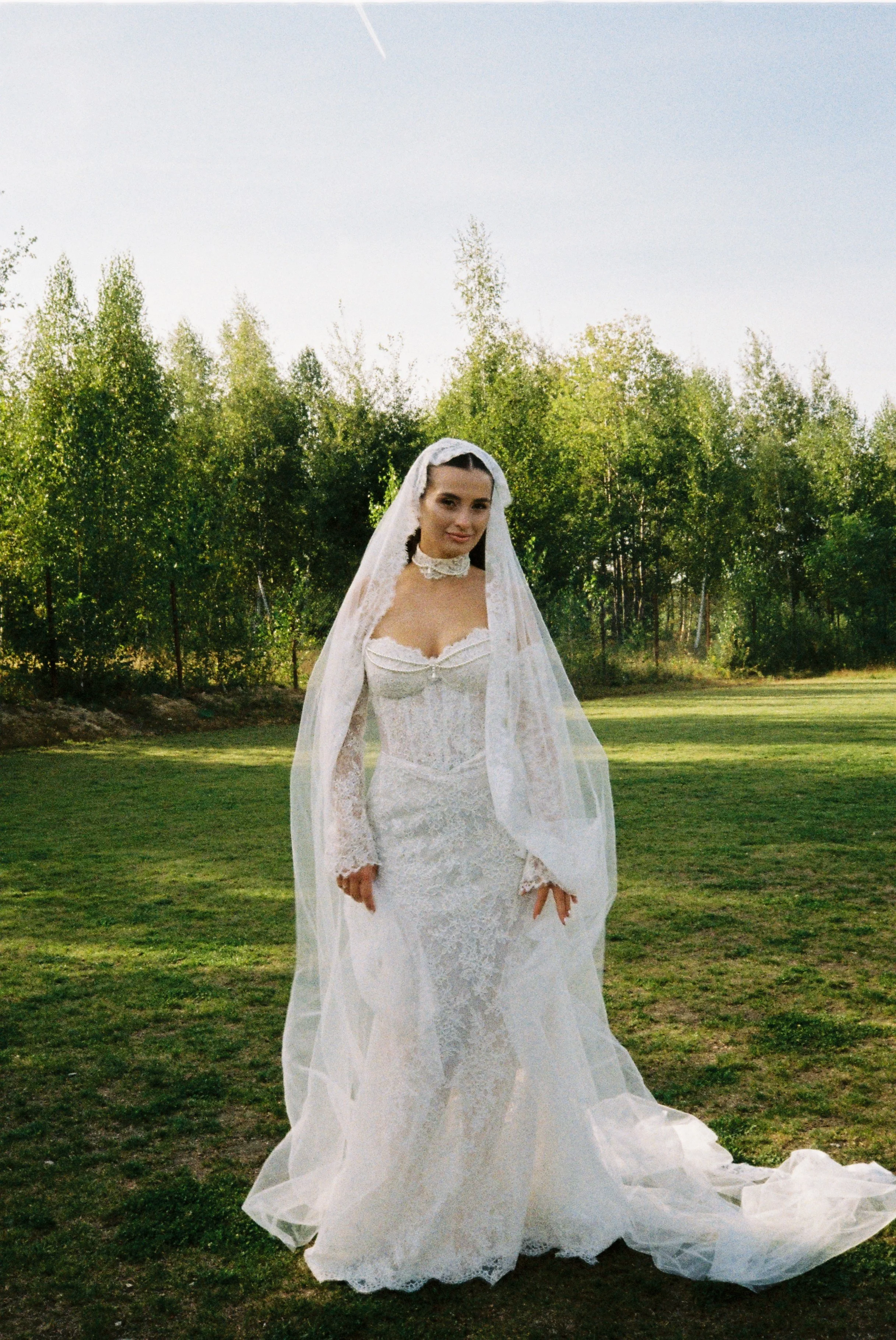 A woman in a white wedding dress and veil stands outdoors on a grassy area with trees in the background, under a clear blue sky.