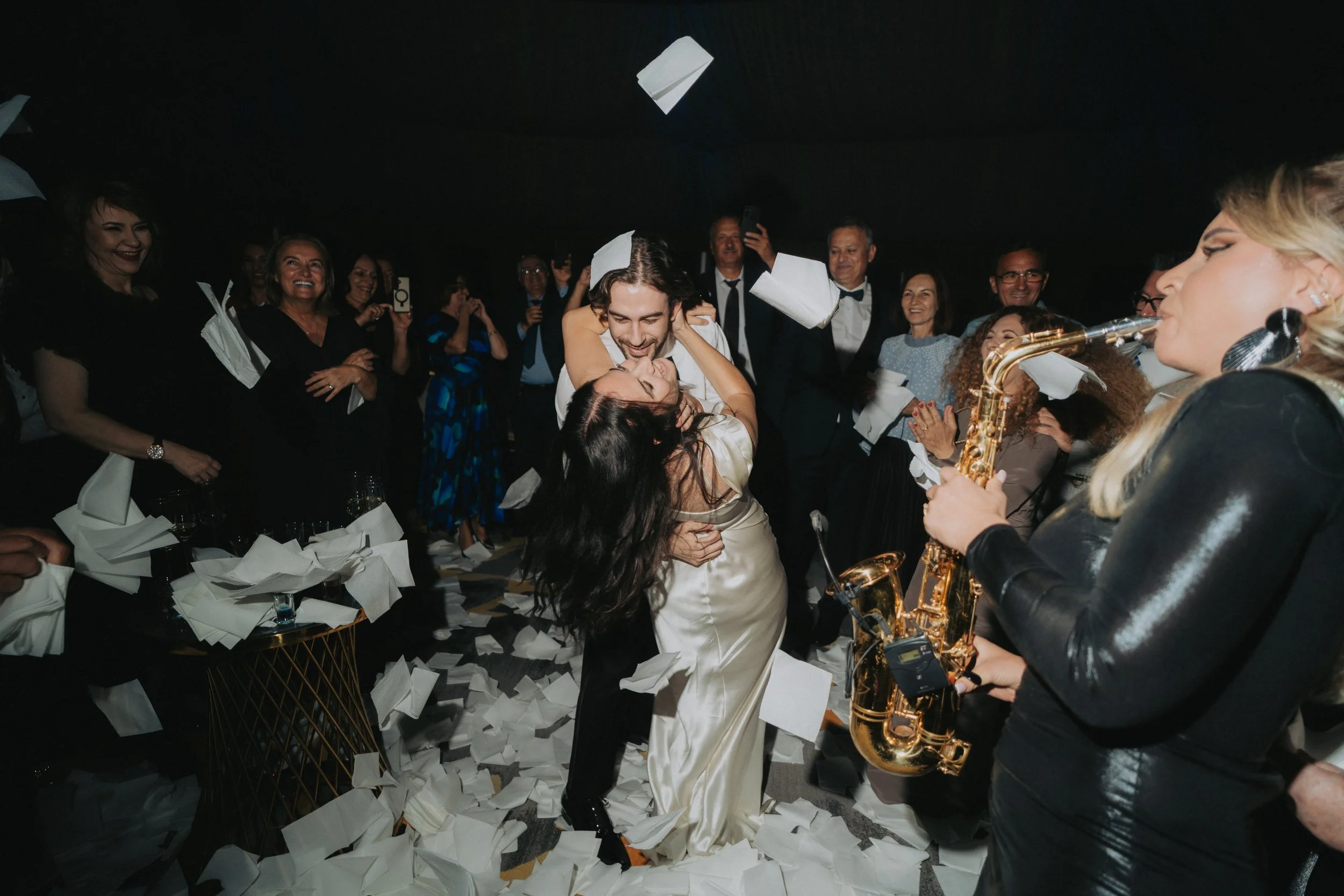Celebrating in a lively party setting, a couple is dancing closely surrounded by joyful guests, with confetti and papers scattered on the floor, and a musician playing the saxophone nearby.