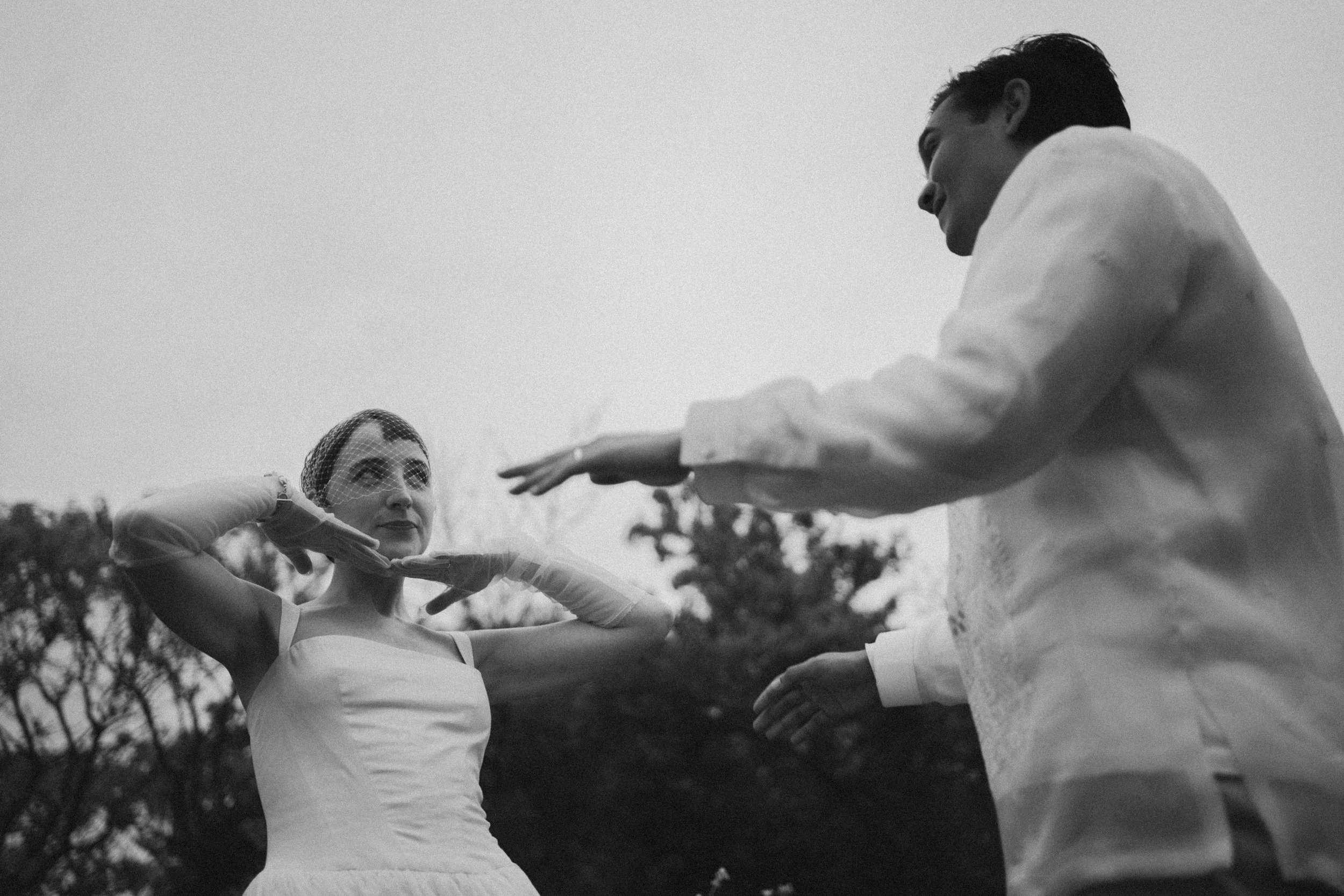 A black and white photo of a man and a woman dancing outdoors, with trees in the background. The woman is wearing a dress and a headband, with her arms raised, and the man is dressed in a suit and holding her hand. They appear to be engaged in a danc