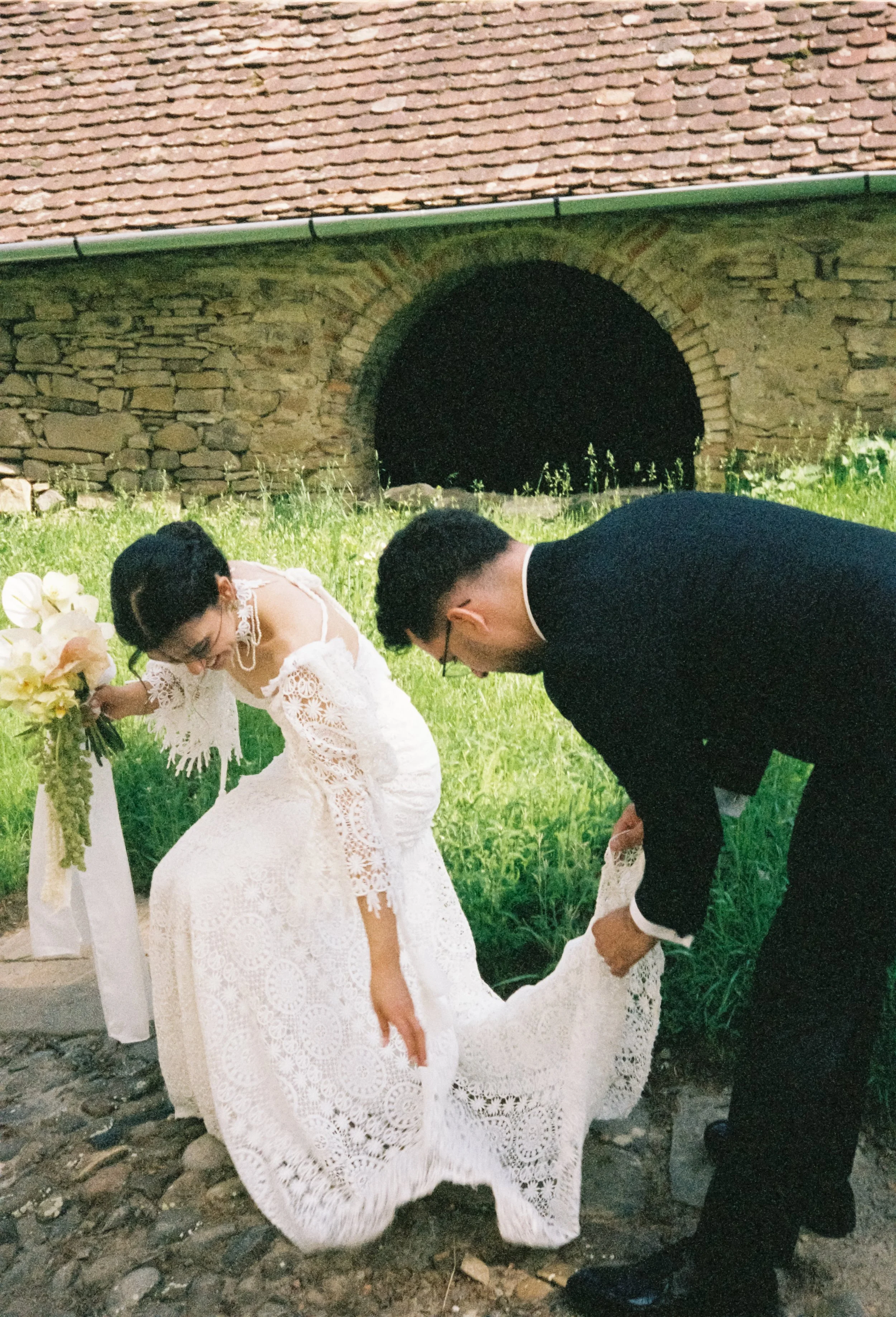 A bride and groom dressed in wedding attire are sharing a moment outside during evening or night. The bride is wearing a white lace dress and the groom is in a black suit, bow tie, and glasses. The groom is helping the bride with her dress. They are 