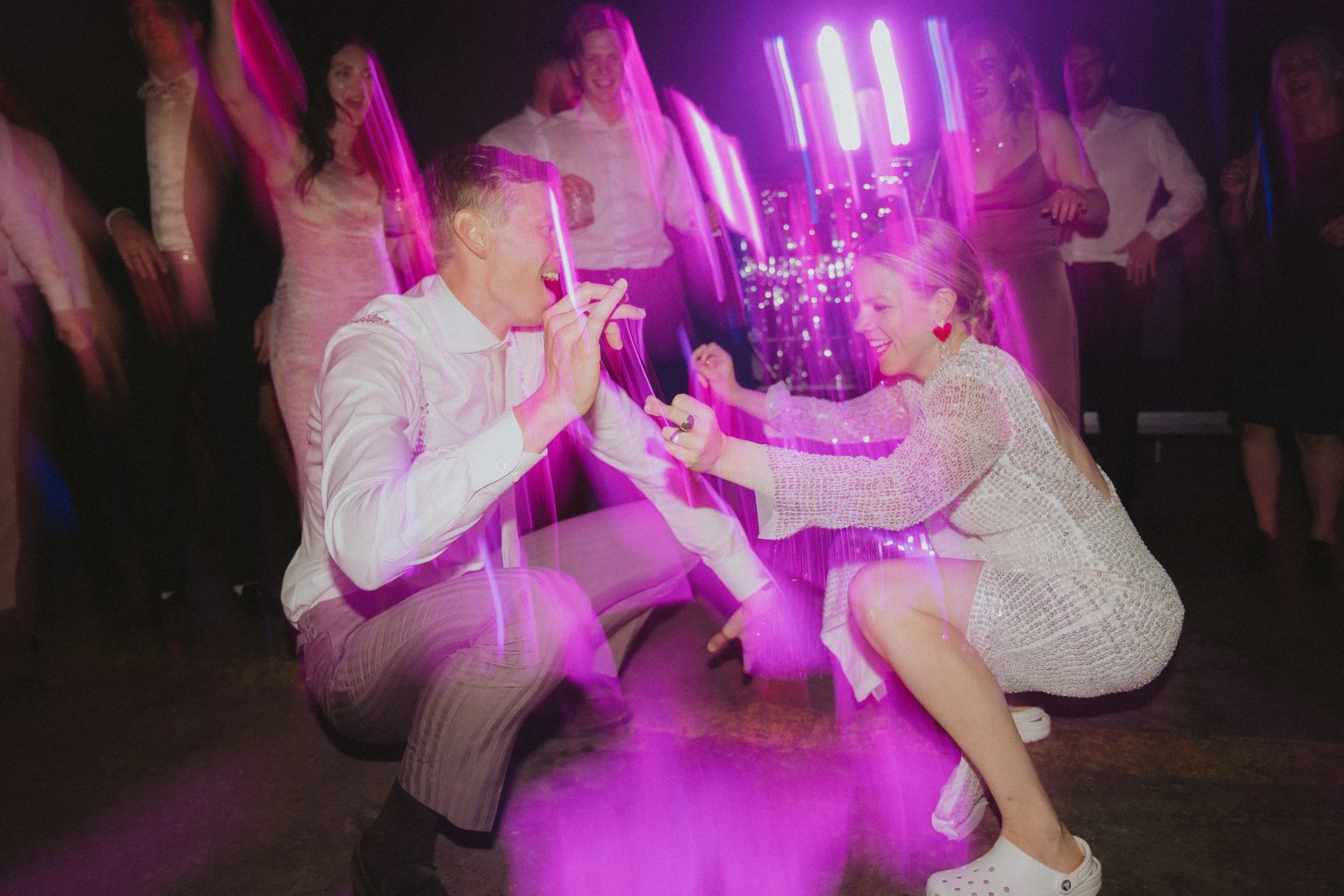 Two people dancing at a party with purple and pink lights, smiling and holding hands.