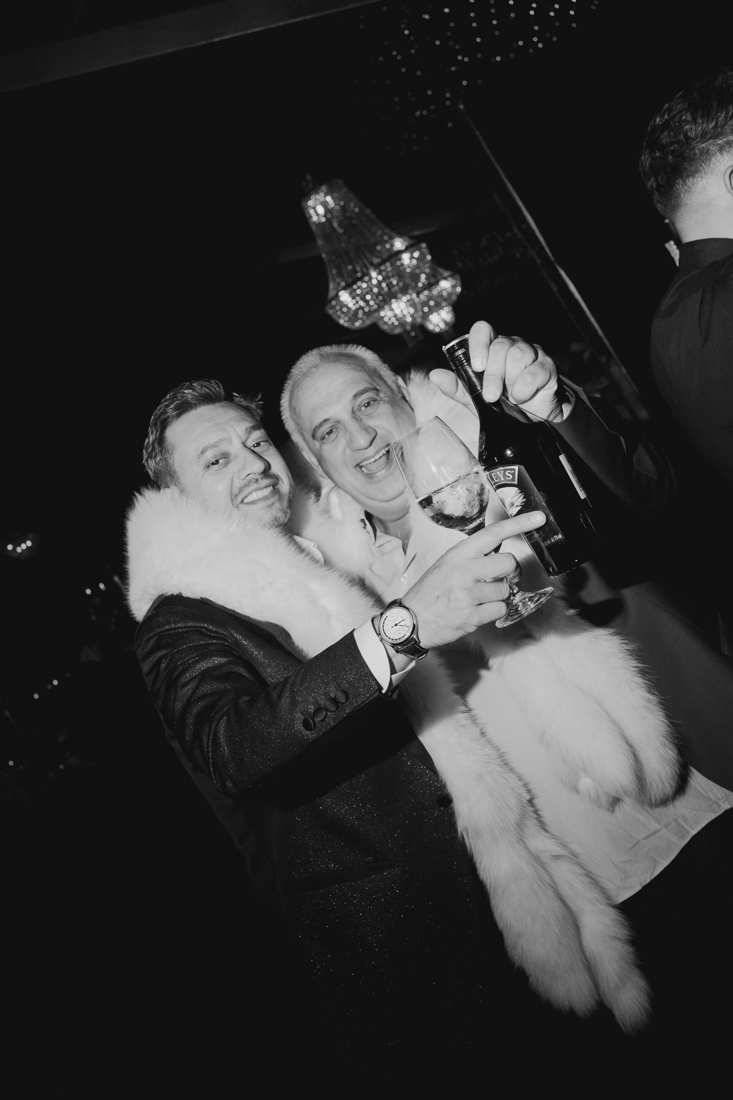 Two men at a celebration, one holding a glass of wine and a bottle, dressed in formal attire with a faux fur coat and a tuxedo with a watch. A chandelier hangs from the ceiling, and a third person with short hair is partially visible on the right.