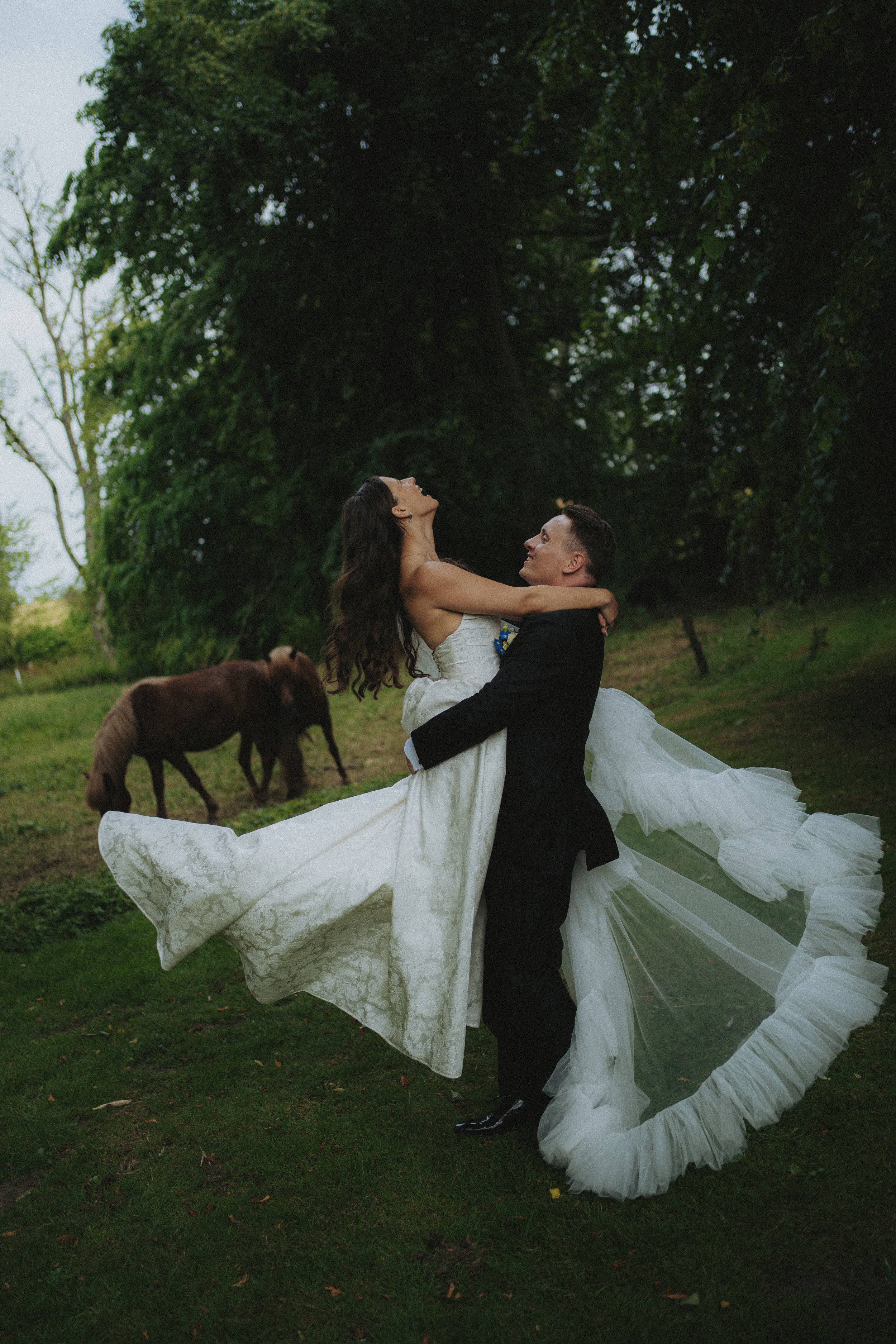A couple dressed in wedding attire dancing outdoors in a natural, wooded setting with two brown dogs nearby.