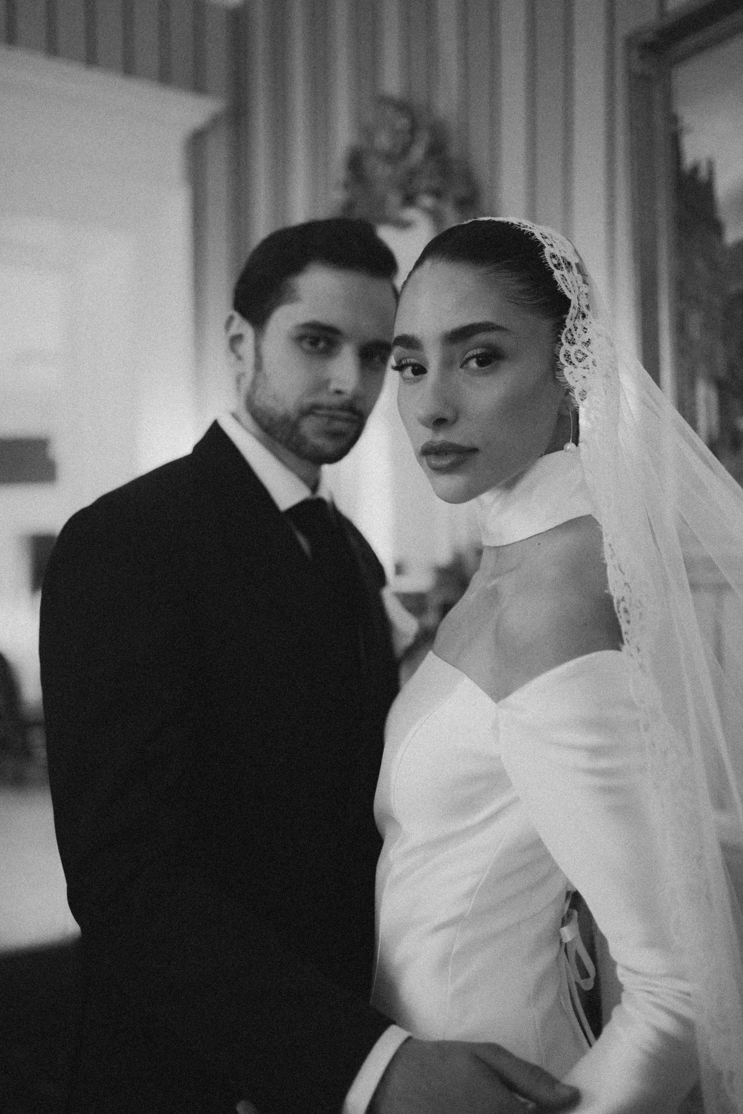 A black and white photograph of a man and woman dressed in formal attire, possibly at a wedding or special event, standing close to each other with serious expressions, in an indoor setting with patterned curtains in the background.