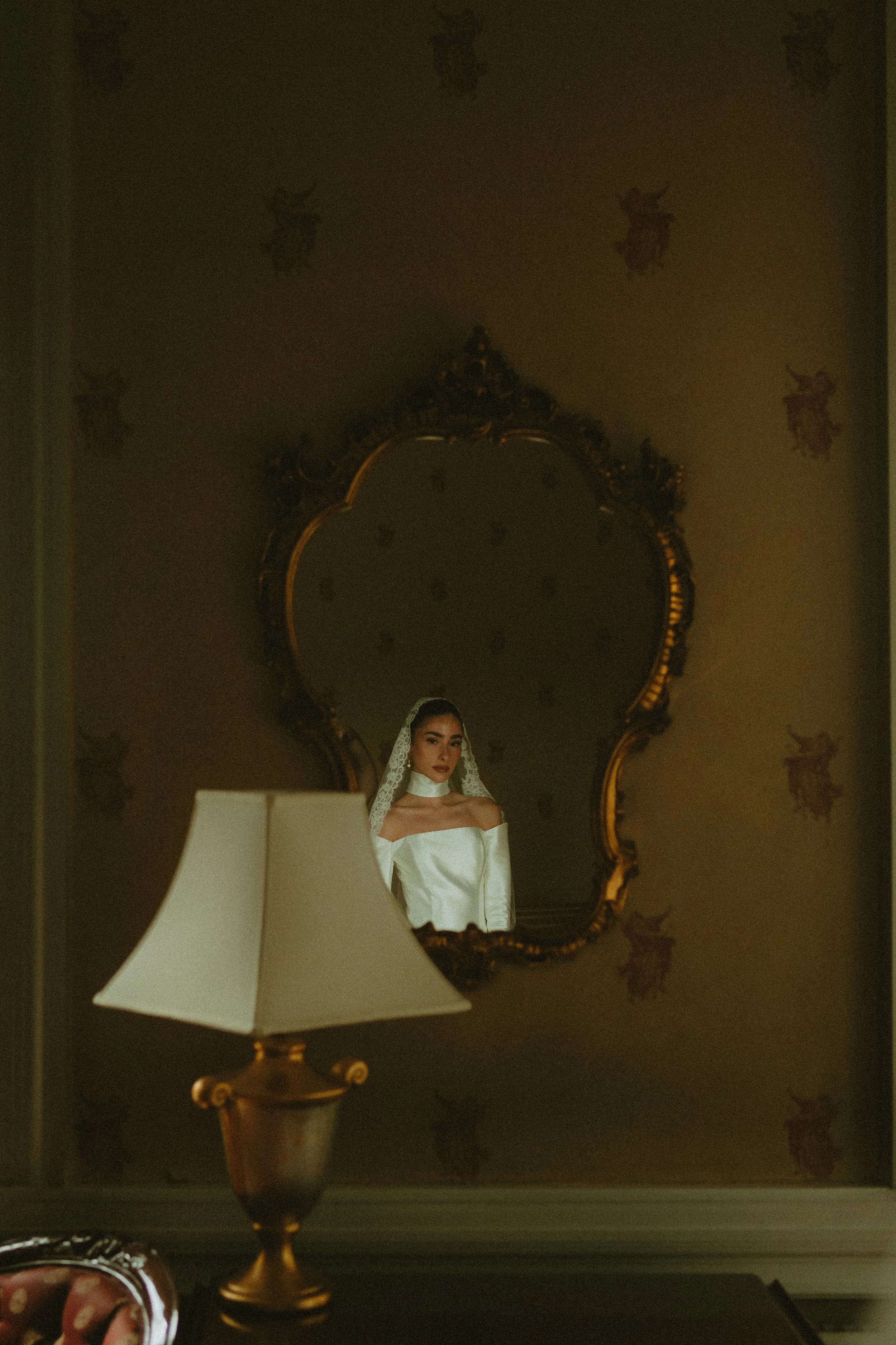 A young woman in a white wedding gown and veil is looking into a mirror in a dimly lit room. The mirror has an ornate gold frame and there is a table lamp with a white lampshade in the foreground.