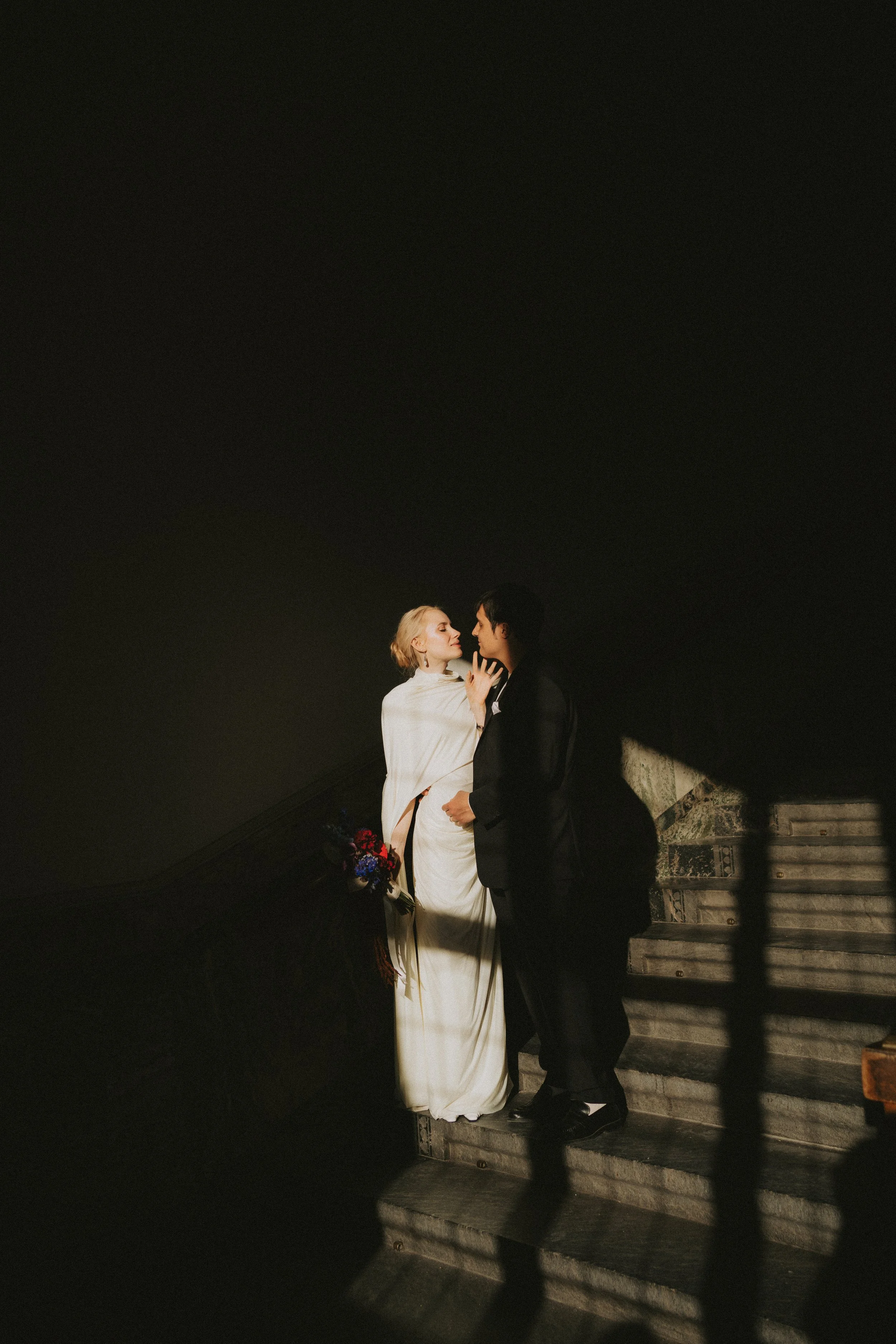 A bride and groom stand on a staircase at night, sharing a tender moment, with the bride holding a bouquet of flowers and wearing a white wedding dress, while the groom is dressed in a dark suit.