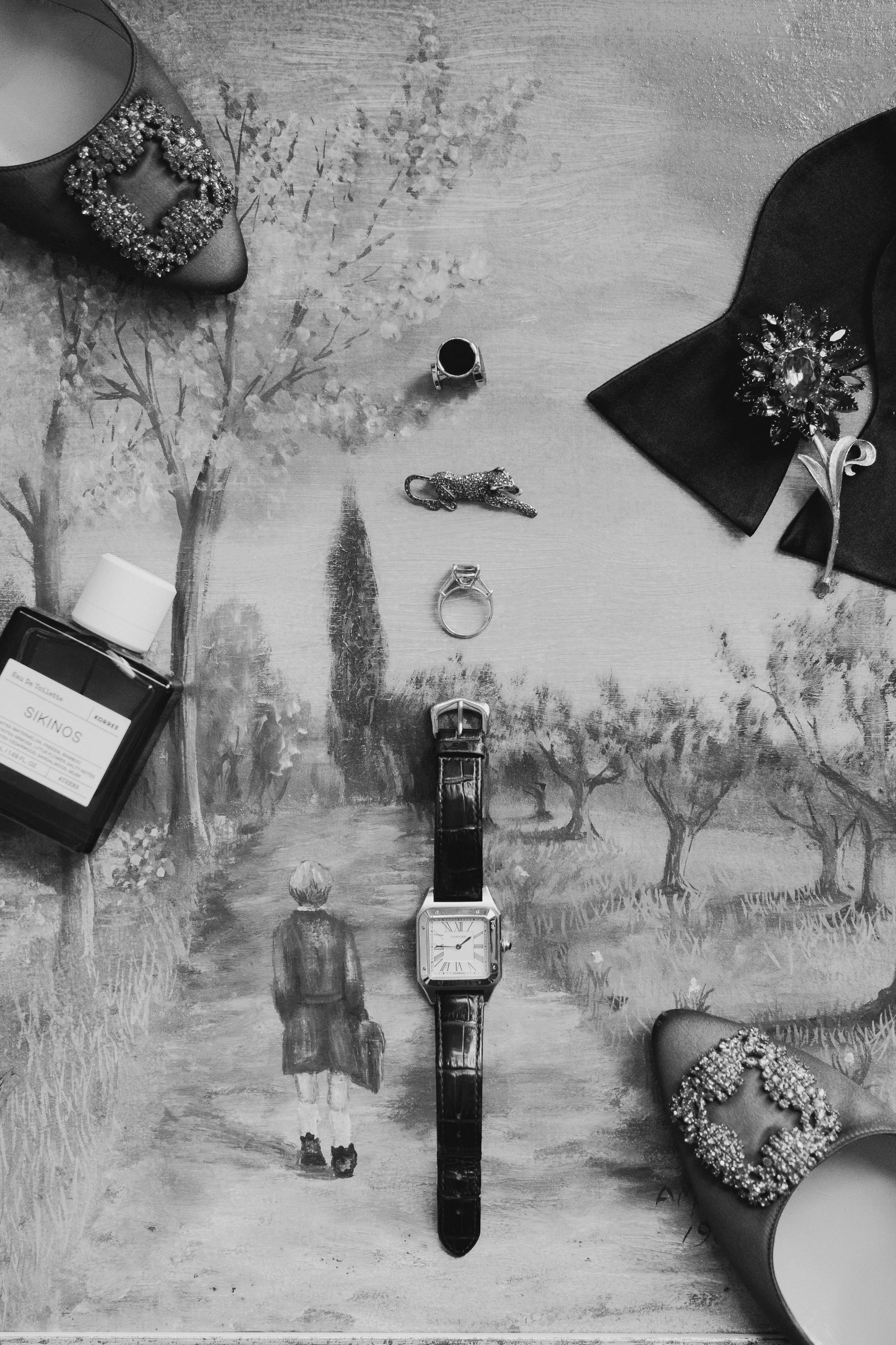 Black-and-white photograph of various jewelry items and a watch on a painted wooden surface. The jewelry includes a pair of embellished shoes, a ring, a brooch, and a small piece of perfume packaging. The surface features a painted landscape with tre