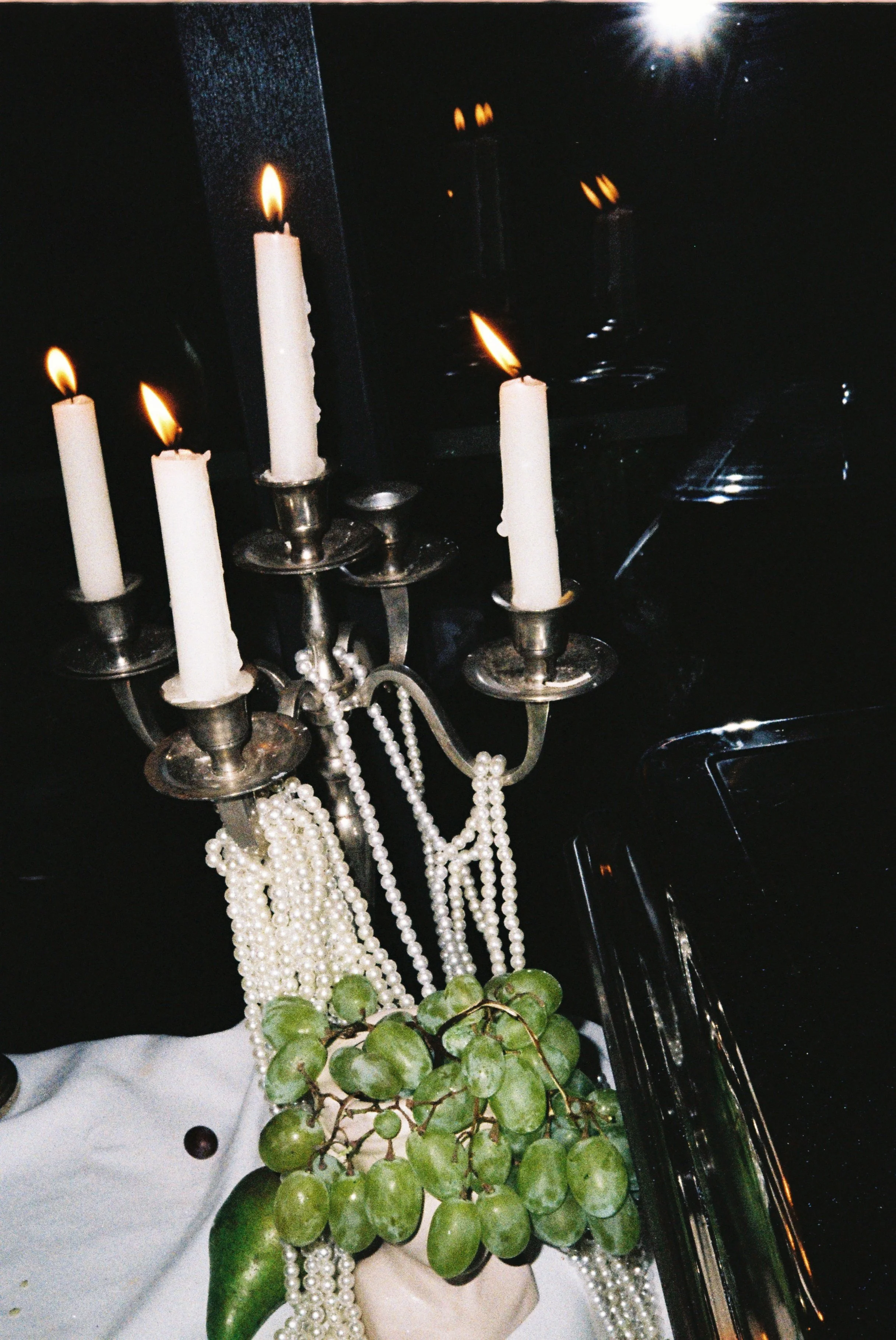 A silver candelabrum with four white lit candles, several strings of pearl necklaces hanging from it, and a small green potted plant with rounded leaves beneath. The background is dark, with a bright light or reflection at the top right and some refl