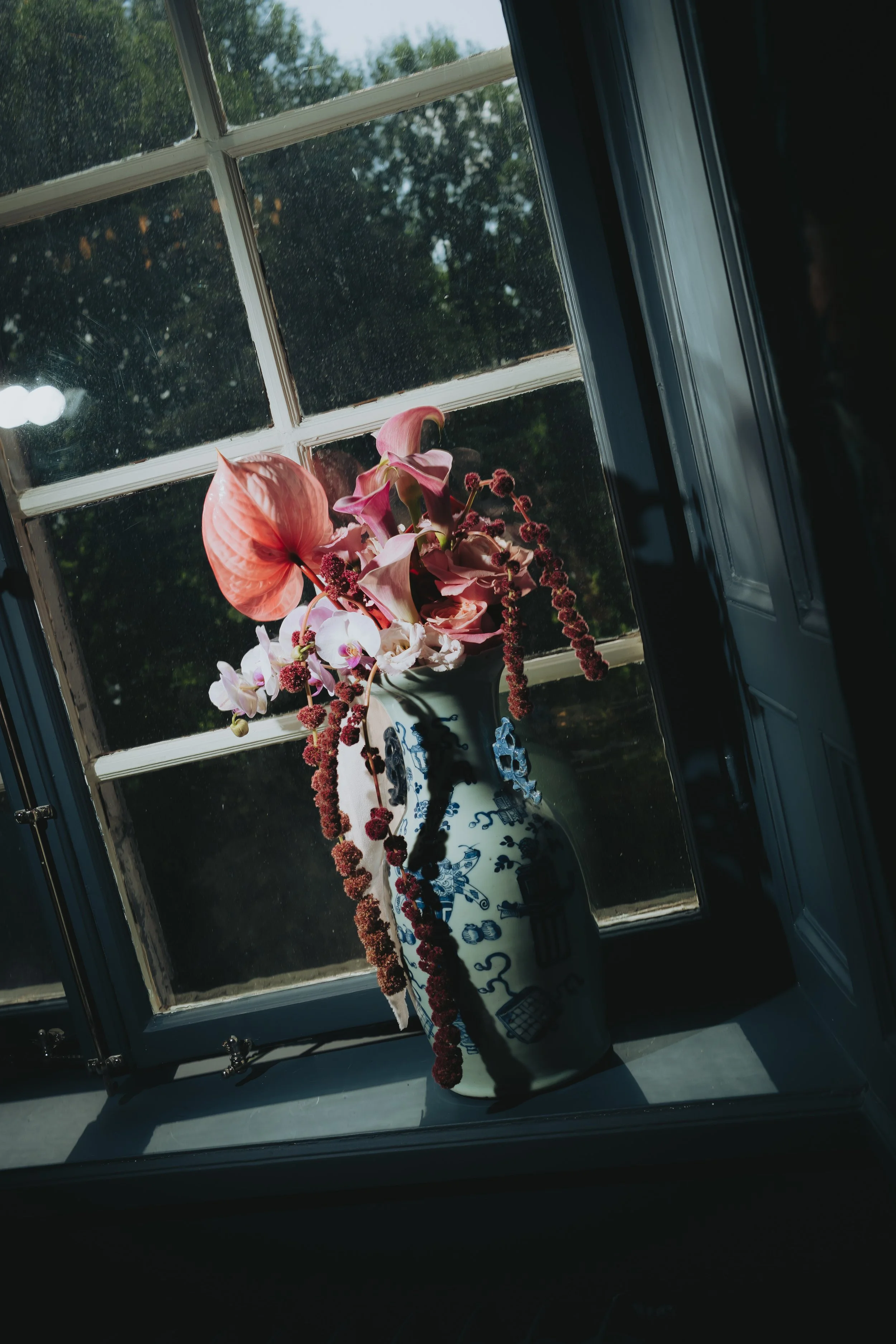 A windowsill with a floral arrangement in a decorative vase. The window is divided into panes, with sunlight shining through, and greenery visible outside.