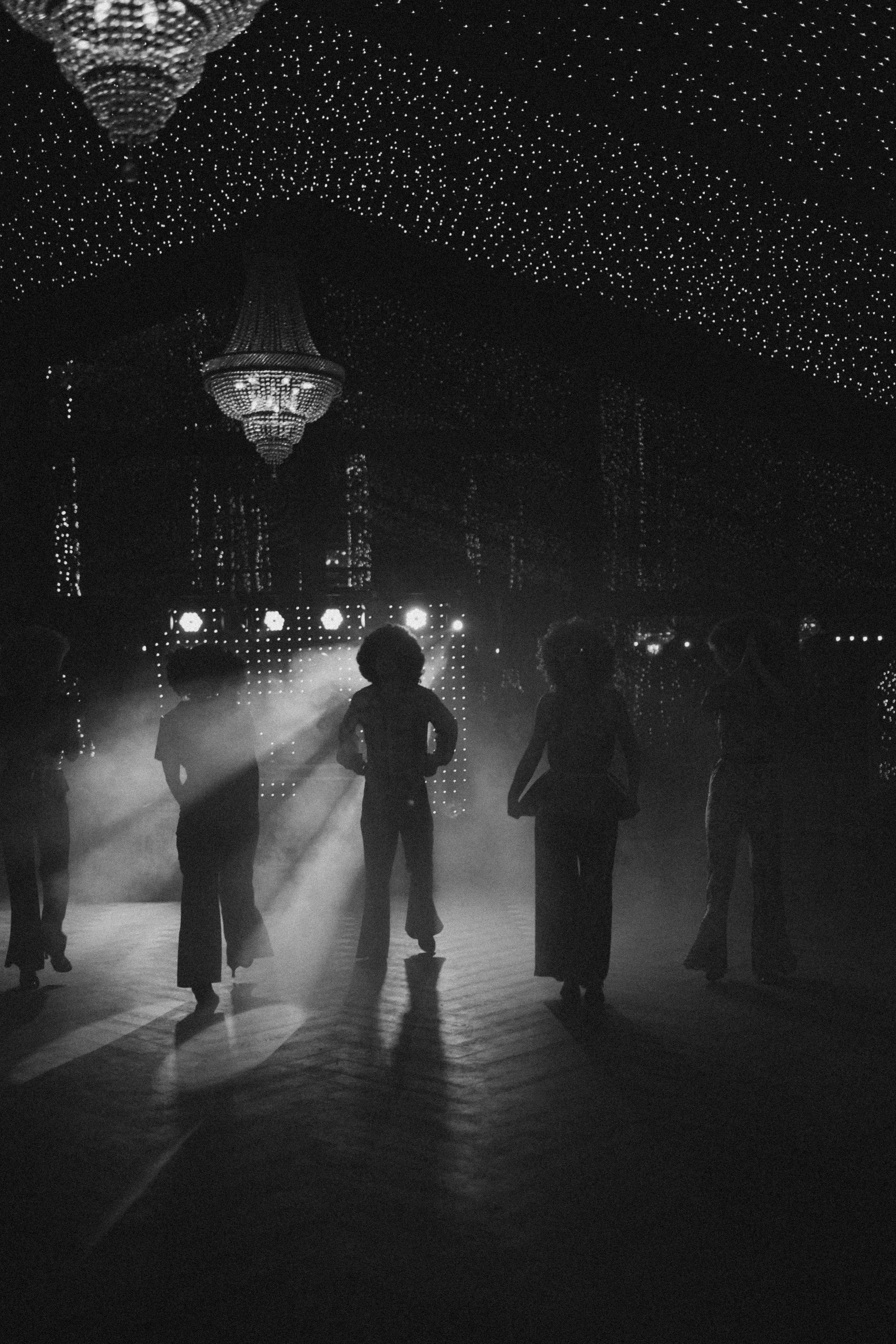 A group of people dancing in a dark, illuminated space with a decorated ceiling and large chandelier, creating a silhouette effect.