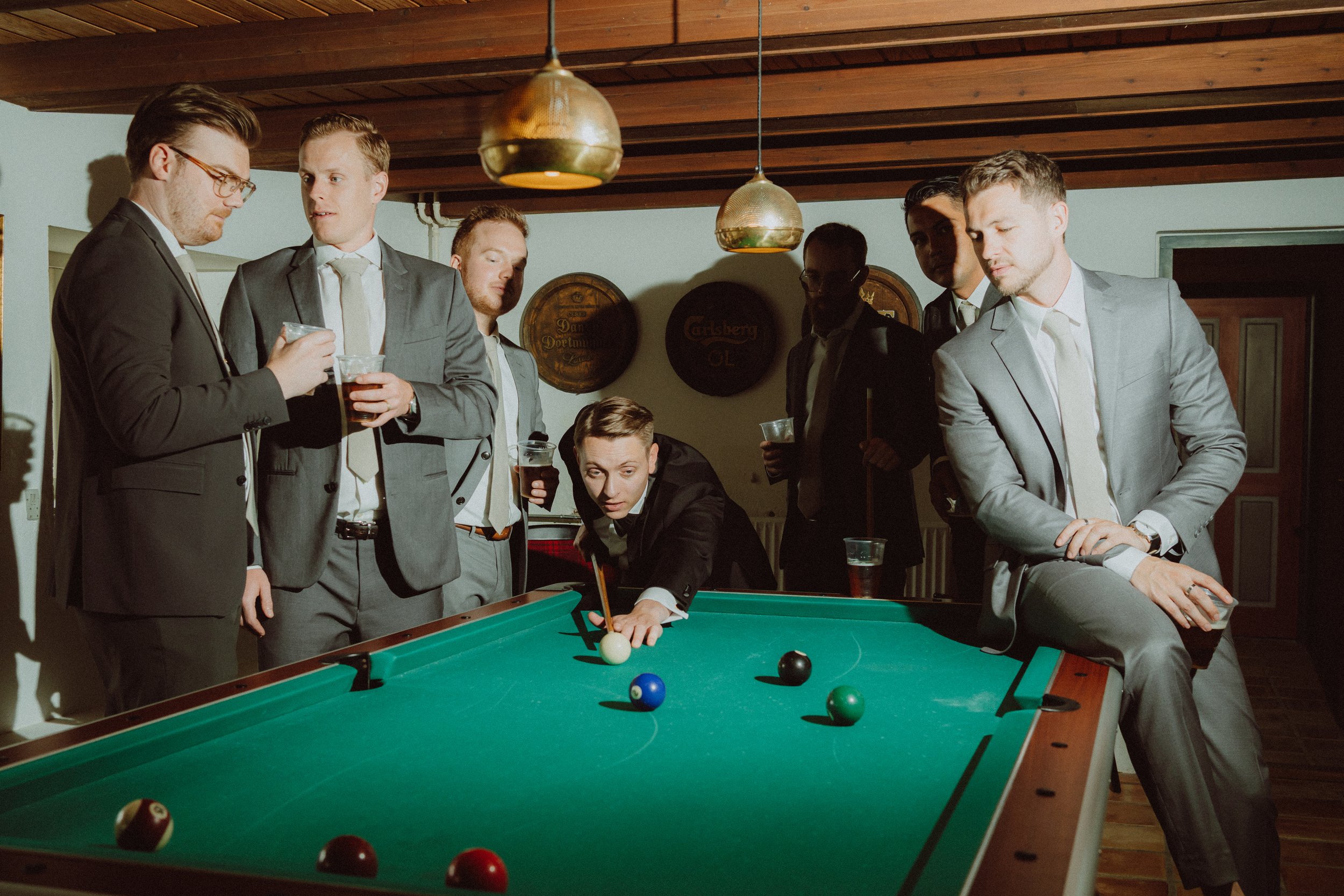 Group of men in suits playing and watching a game of pool in a room with wood-paneled ceiling, gold pendant lights, and decorative wall plaques.