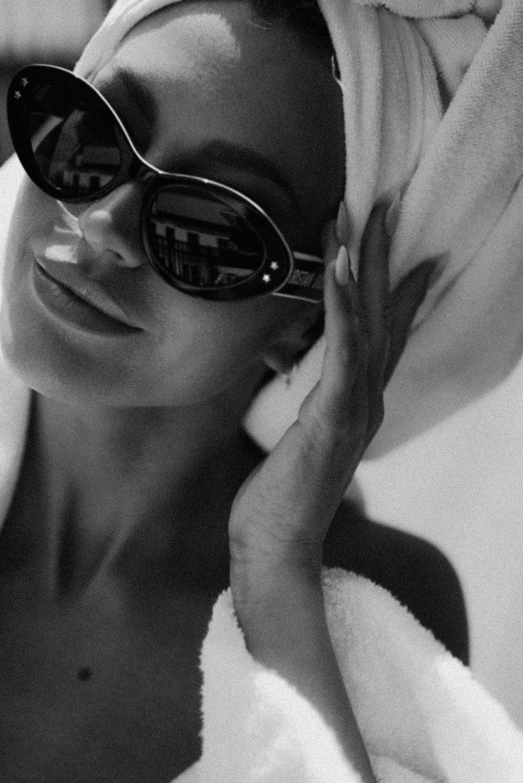 A woman with dark hair wearing sunglasses and a white bathrobe, resting her face and head against her hand.