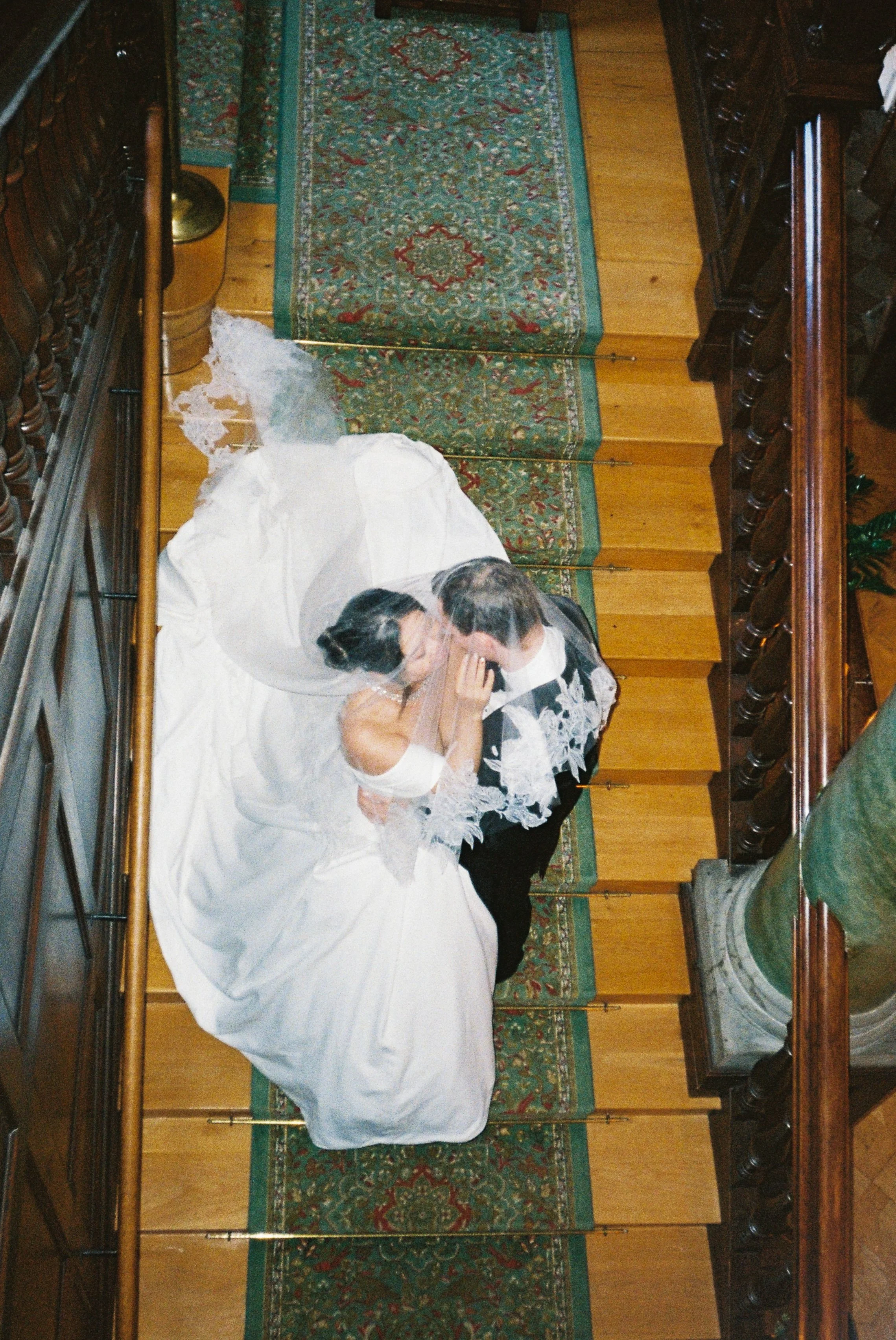 A bride and groom sharing a kiss under a sheer veil in a rustic indoor setting with wooden floors, an ornate rug, and wooden furniture on the sides.
