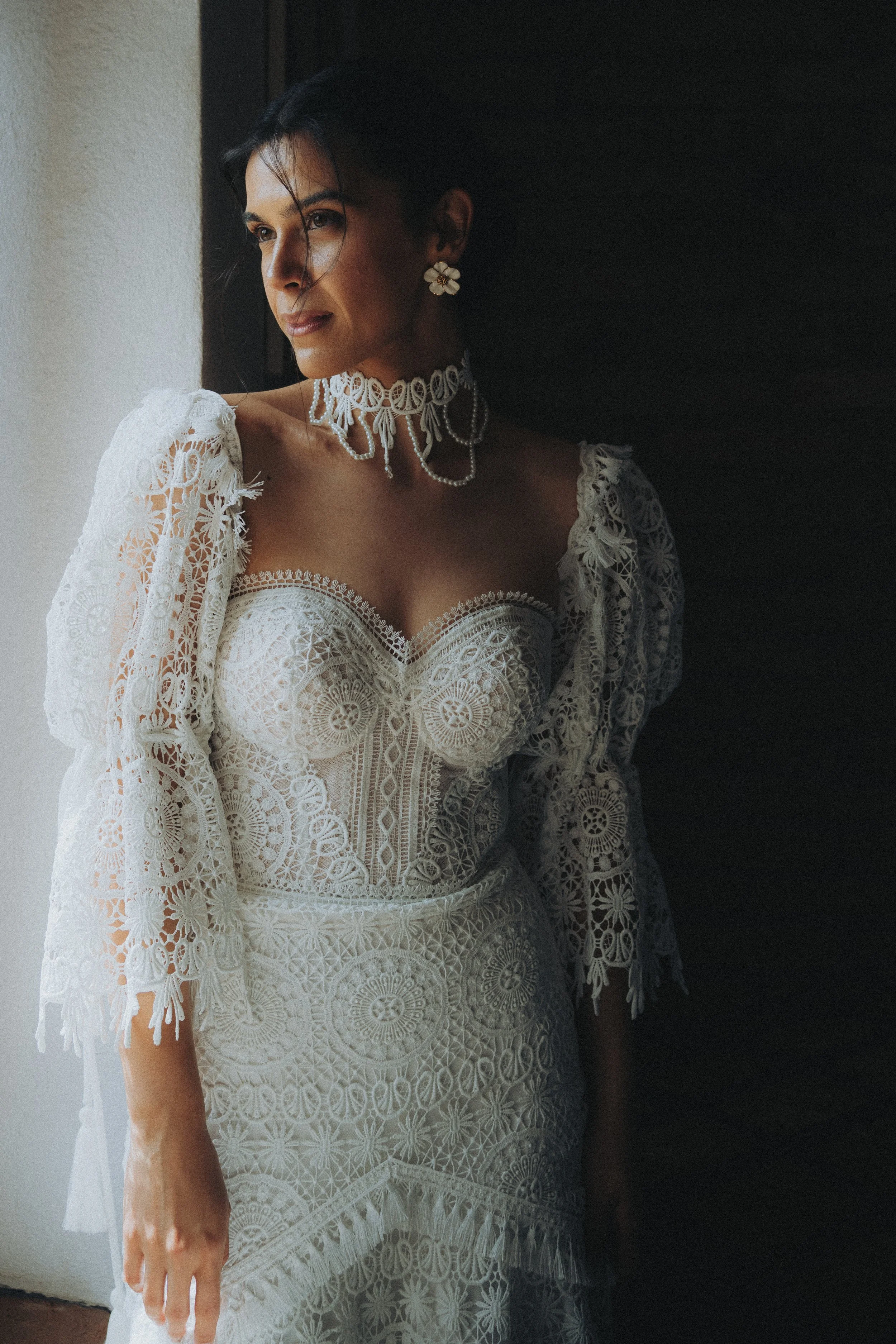 Woman wearing a white lace dress and vintage jewelry, standing near a window, with soft lighting illuminating her face.