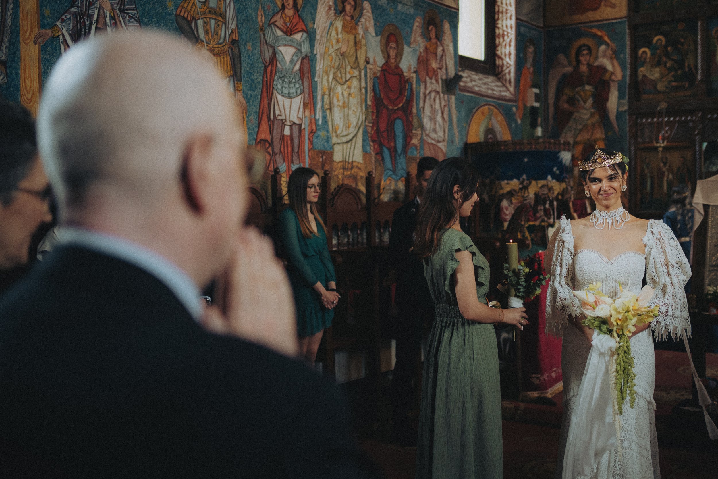 A woman wearing a white lace dress and a crown holds a bouquet of flowers during a wedding ceremony in a church with colorful religious murals.